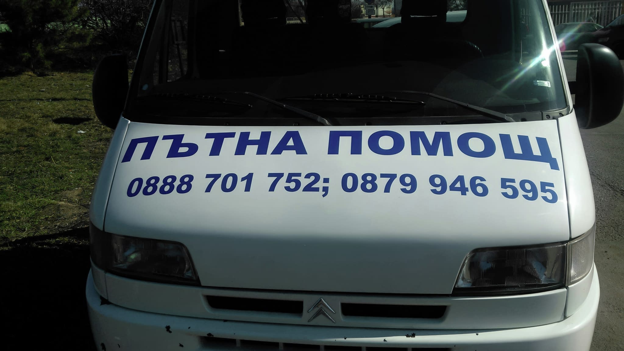 Бял бус с надпис Пътна помощ и телефони за връзка, брандиран на предния капак с големи сини букви.