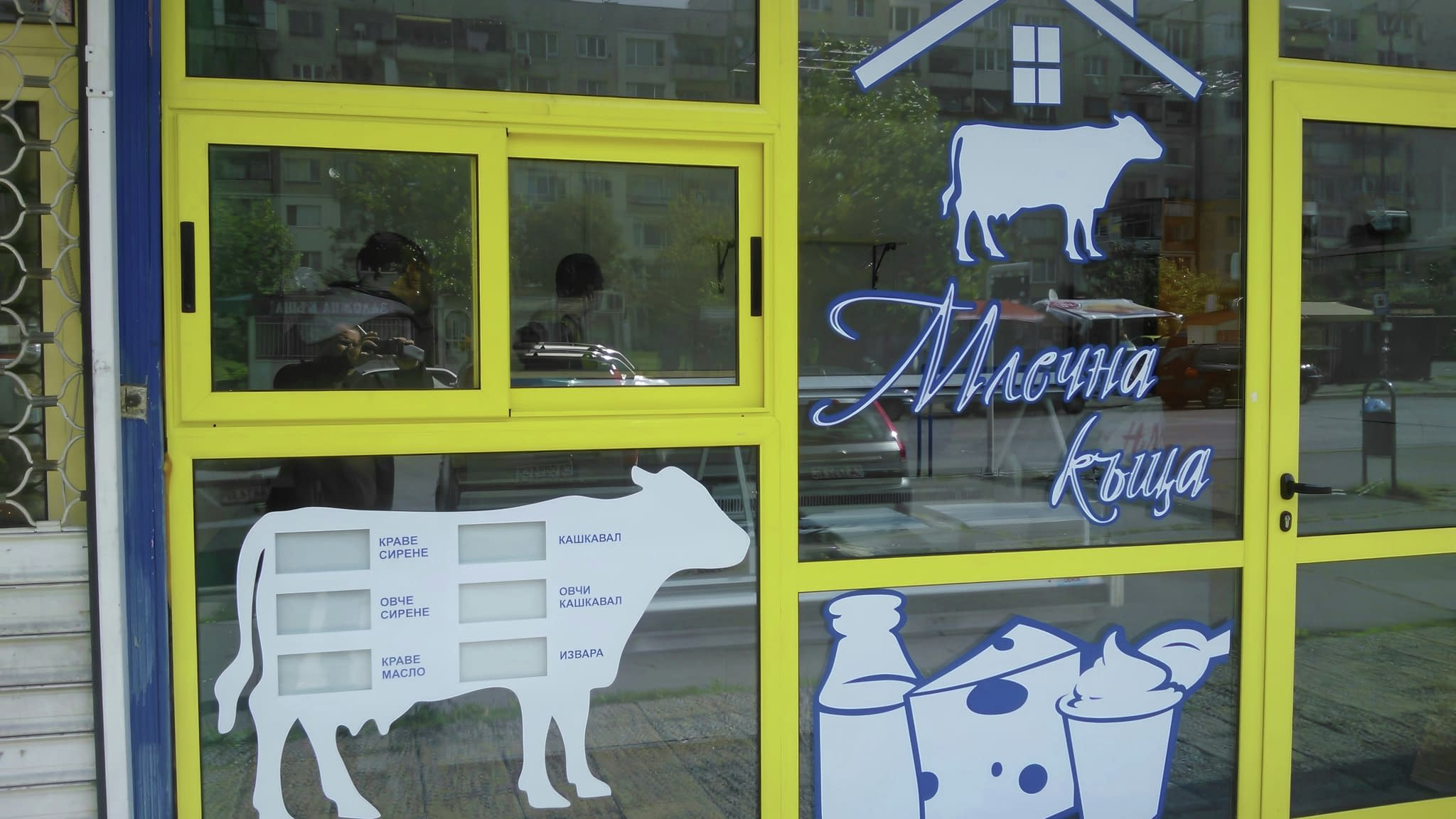 Витрина на млечен магазин с жълта рамка, украсена с бели фолио стикери на продукти и крави.