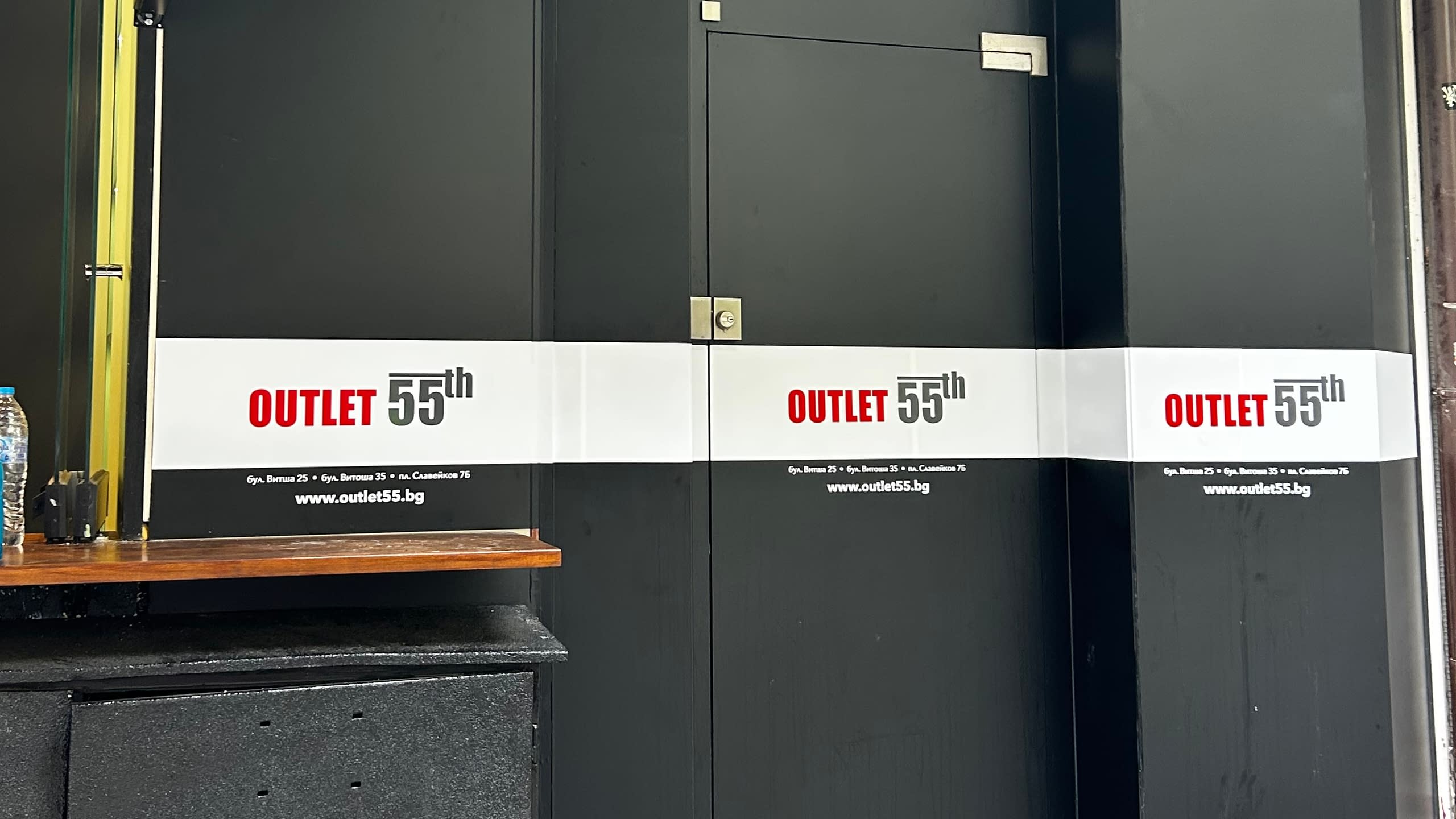 Тъмна фасада на магазин "OUTLET 55th", фолирана с бяла лента с логото, адреса и уебсайта на фирмата.