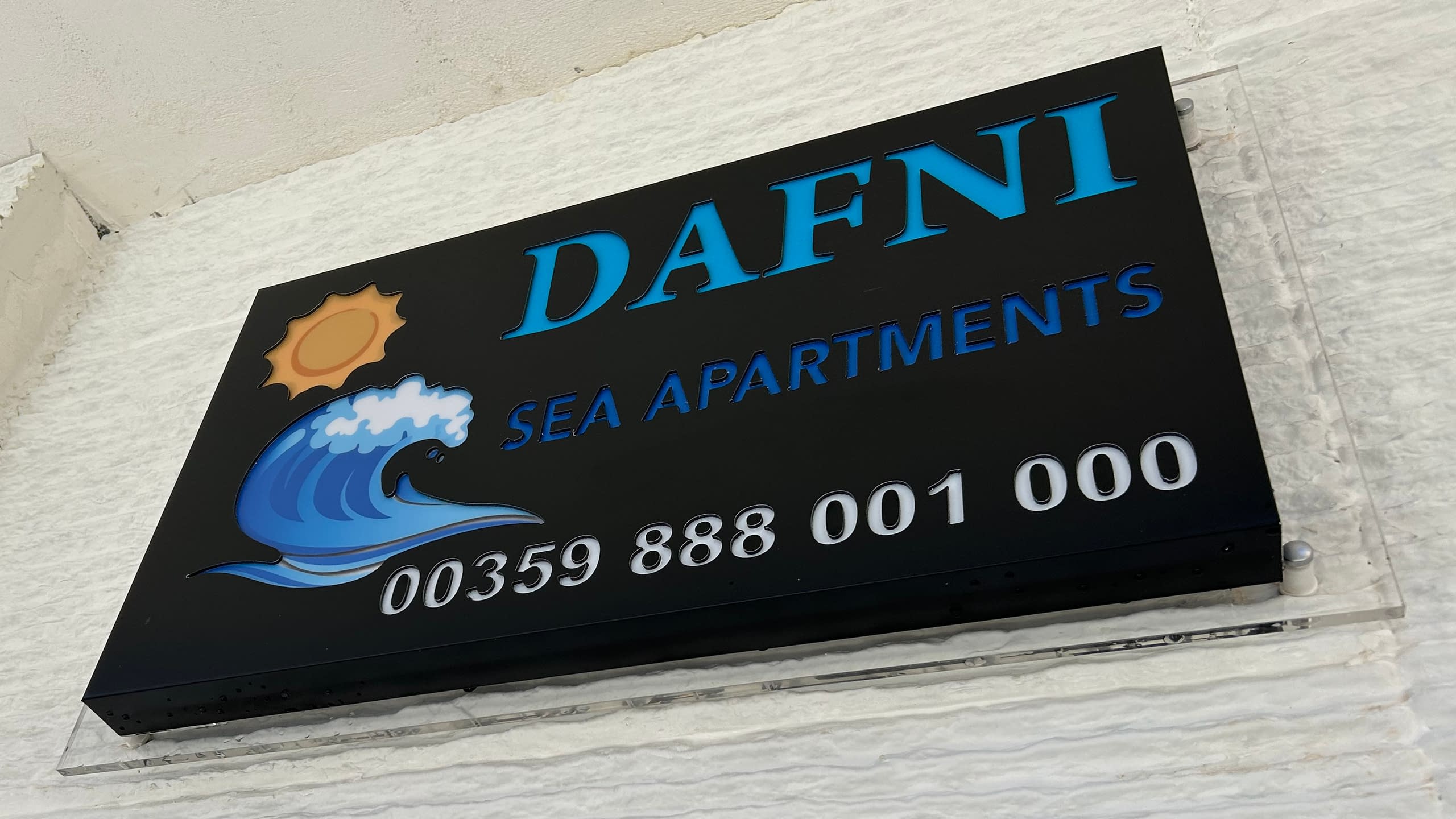 Интериорна светеща табела за "Dafni Sea Apartments" с лого на вълна и слънце, монтирана на плексигласова основа.