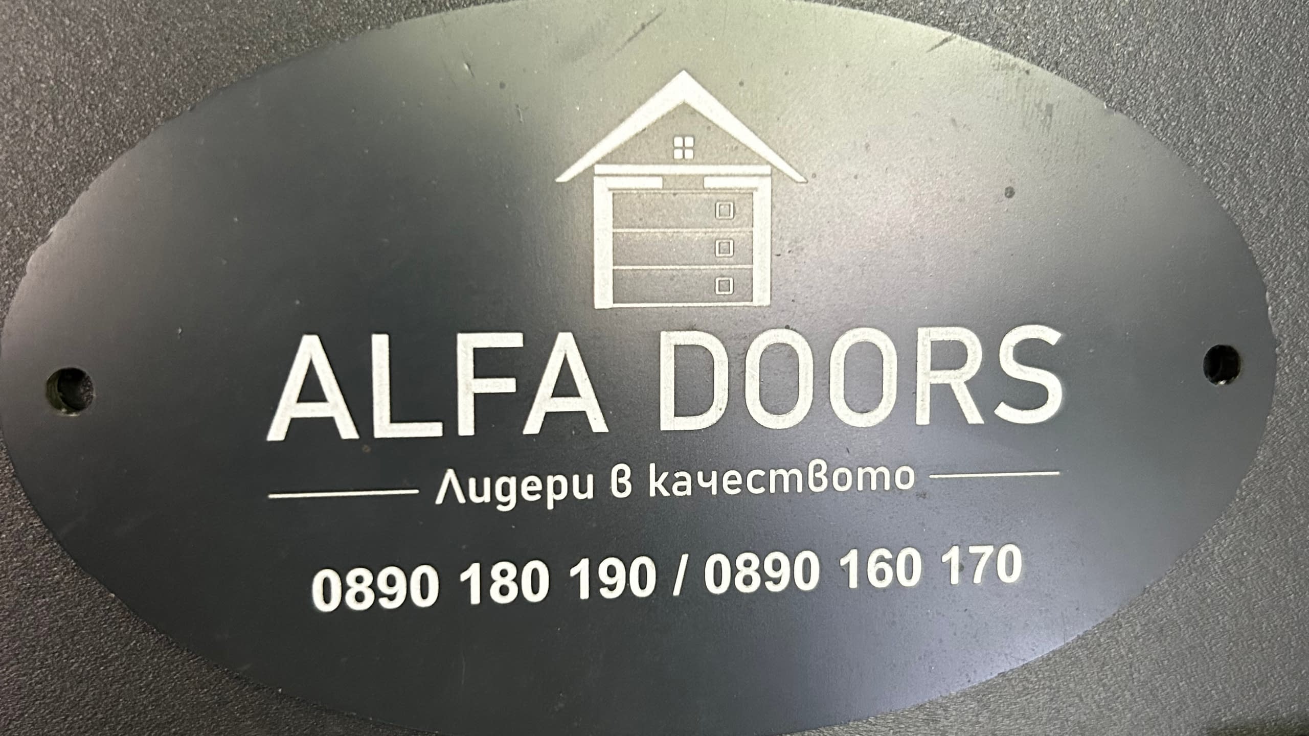 Овална гравирана табела от черен материал за фирма "Alfa Doors" с лого, слоган и телефонни номера.