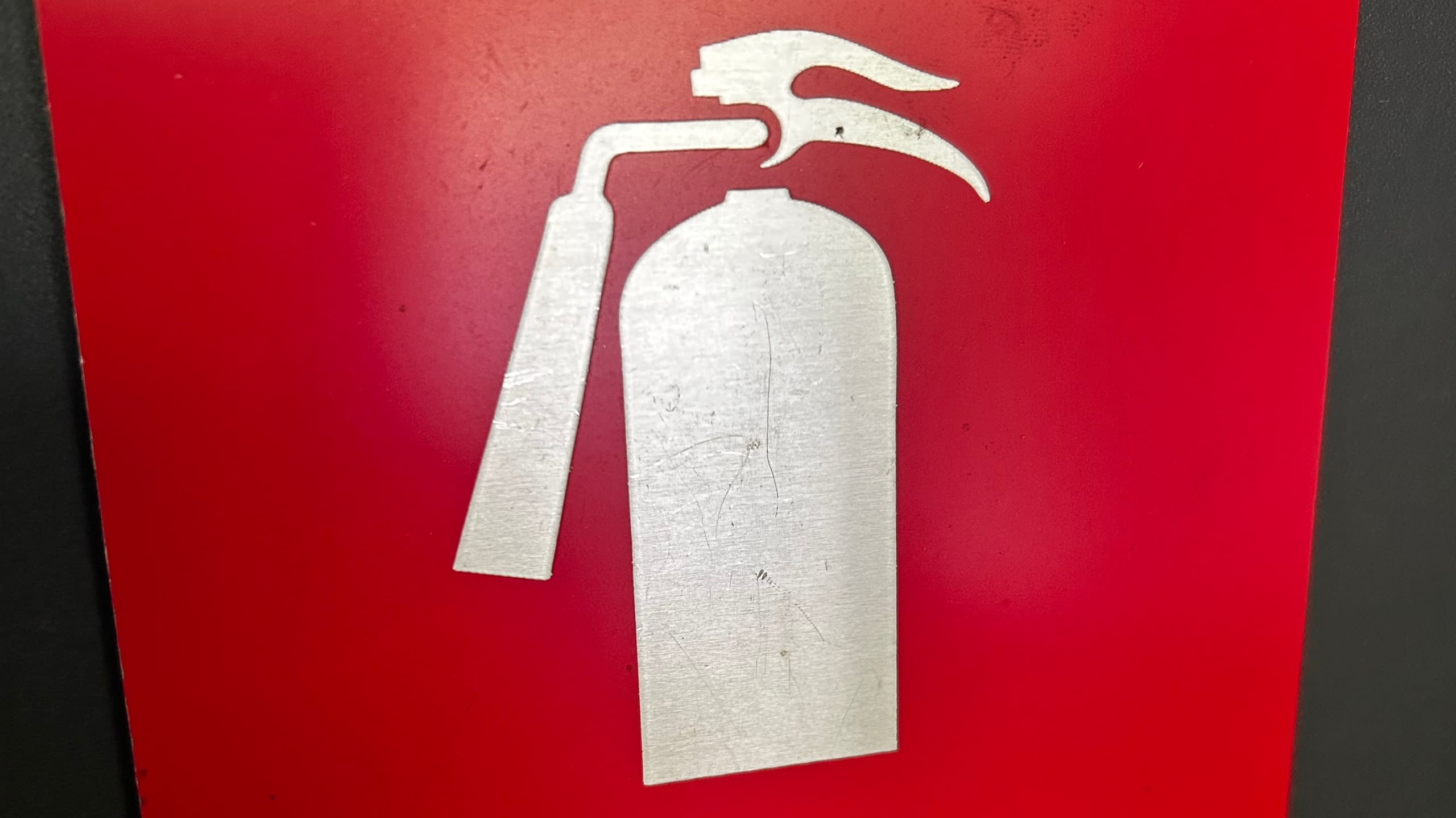 Указателна интериорна табела за пожарогасител, изработена от червена основа и метален сребрист символ.