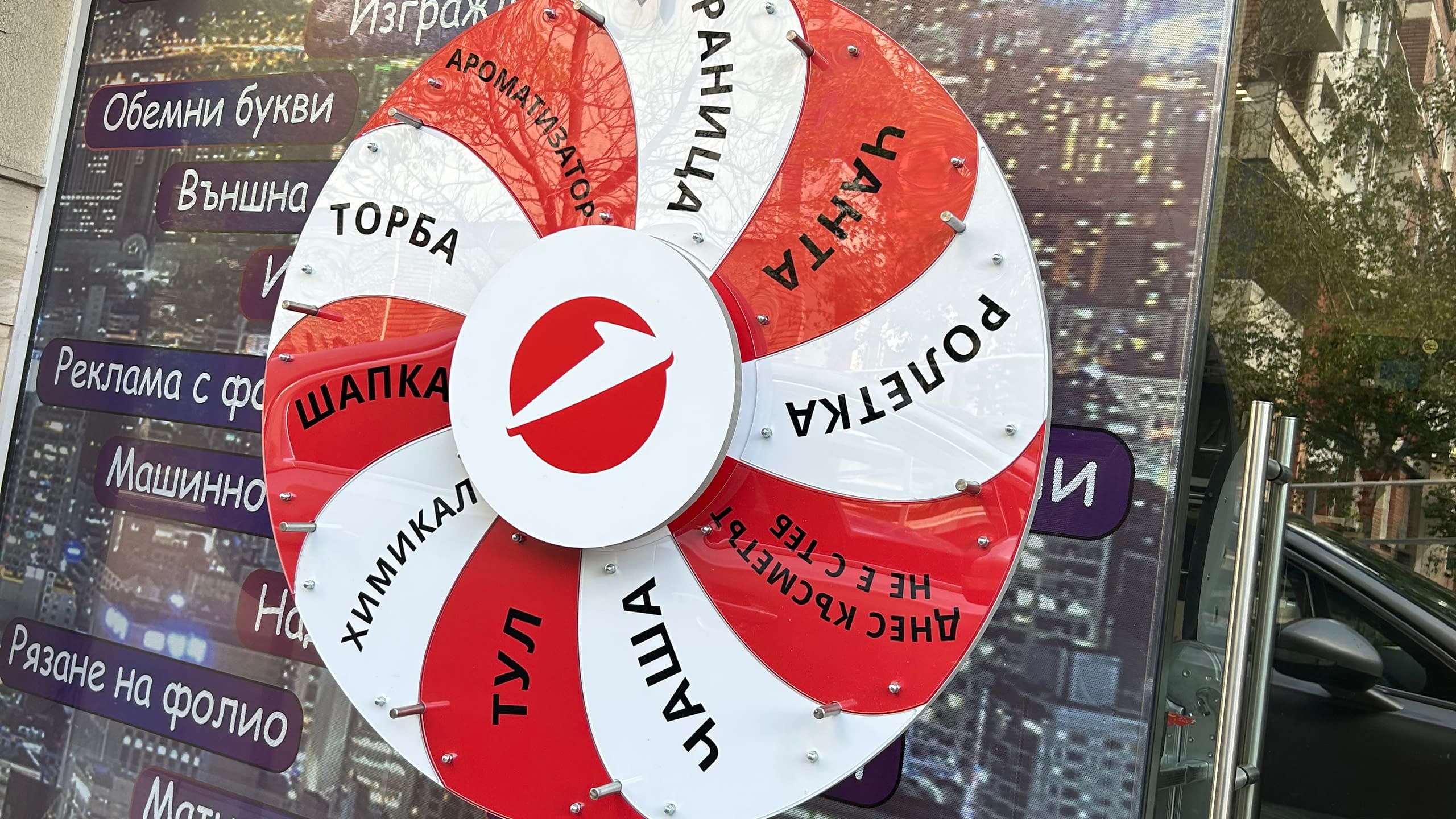 Изработка на рекламно колело на късмета в червено, бяло и сиво, със сектори за различни предметни награди като "Шапка", "Раница" и "Химикал" за промоционална игра.