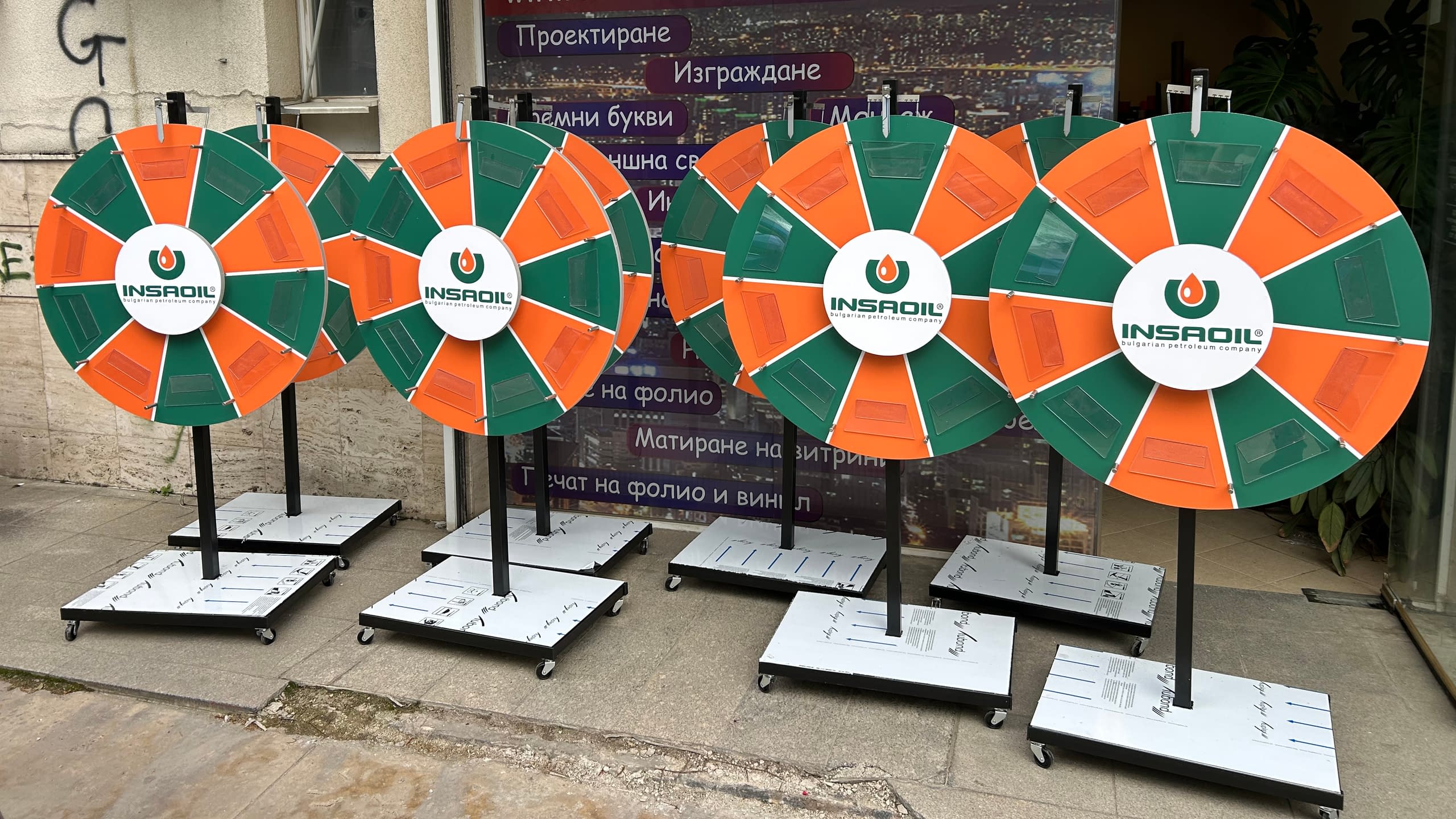 Множество еднакви колела на късмета, брандирани за бензиностанции INSADIL в оранжево и зелено, поставени на стабилни метални стойки с колелца.