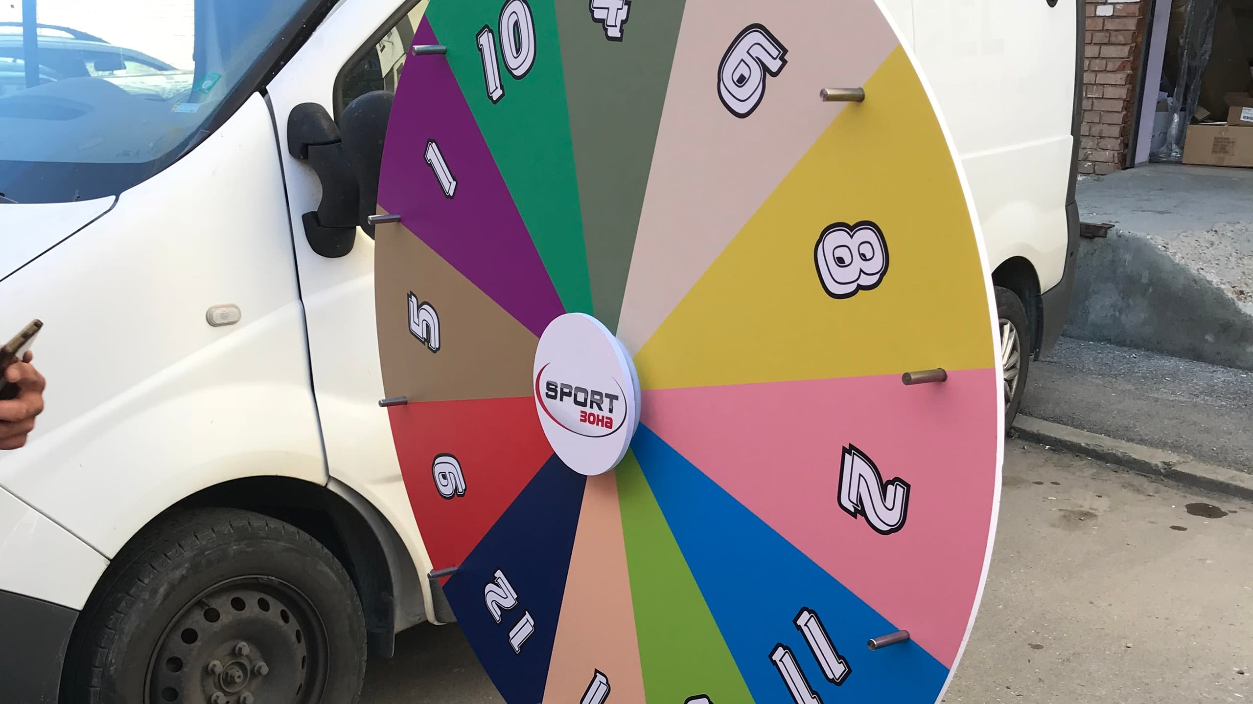 Голямо, многоцветно колело на късмета, брандирано за "SPORT ZONA", със сектори с различни числа. Колелото е поставено на открито до бял микробус.