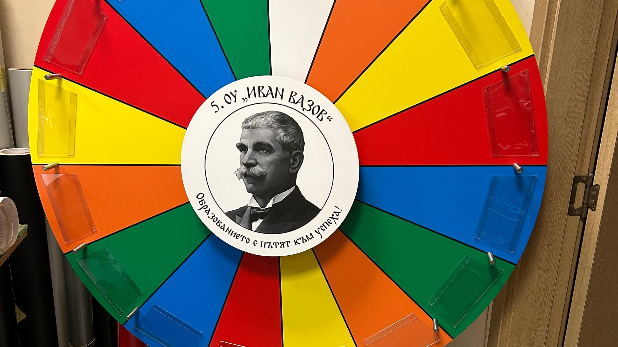 Образователно колело на късмета, изработено за 5.ОУ "Иван Вазов", с портрет на патрона в центъра, цветни сектори и прозрачни джобове за поставяне на сменяеми награди или въпроси.