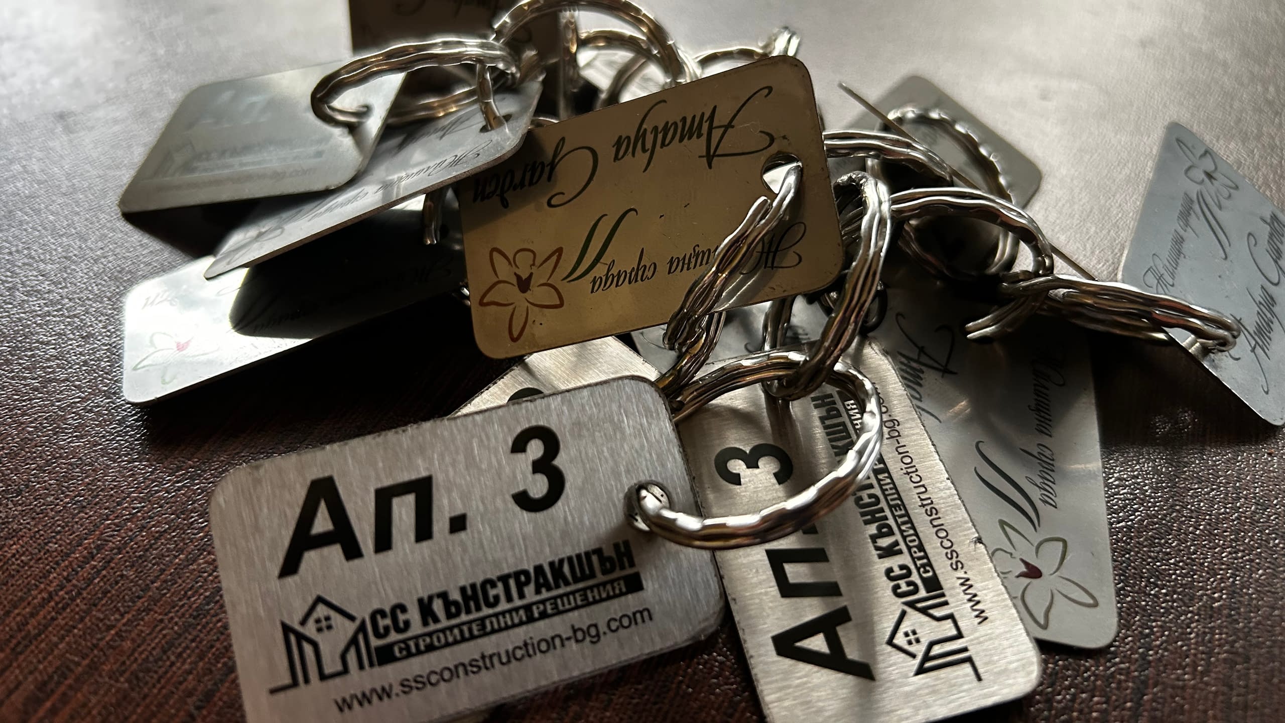 Купчина метални ключодържатели с лазерно гравиране - едните са брандирани за строителна фирма "СС Кънстракшън", а други са с декоративен надпис и флорални мотиви.
