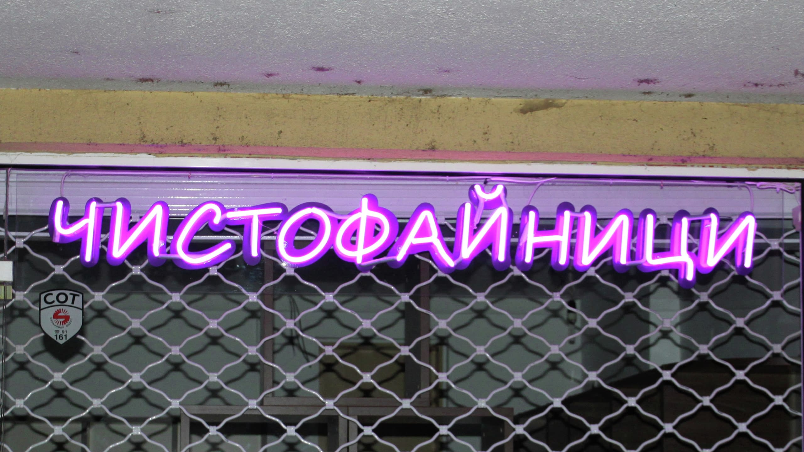 Лилав неонов надпис "Чистофайници", монтиран на витрина зад охранителна метална щора.