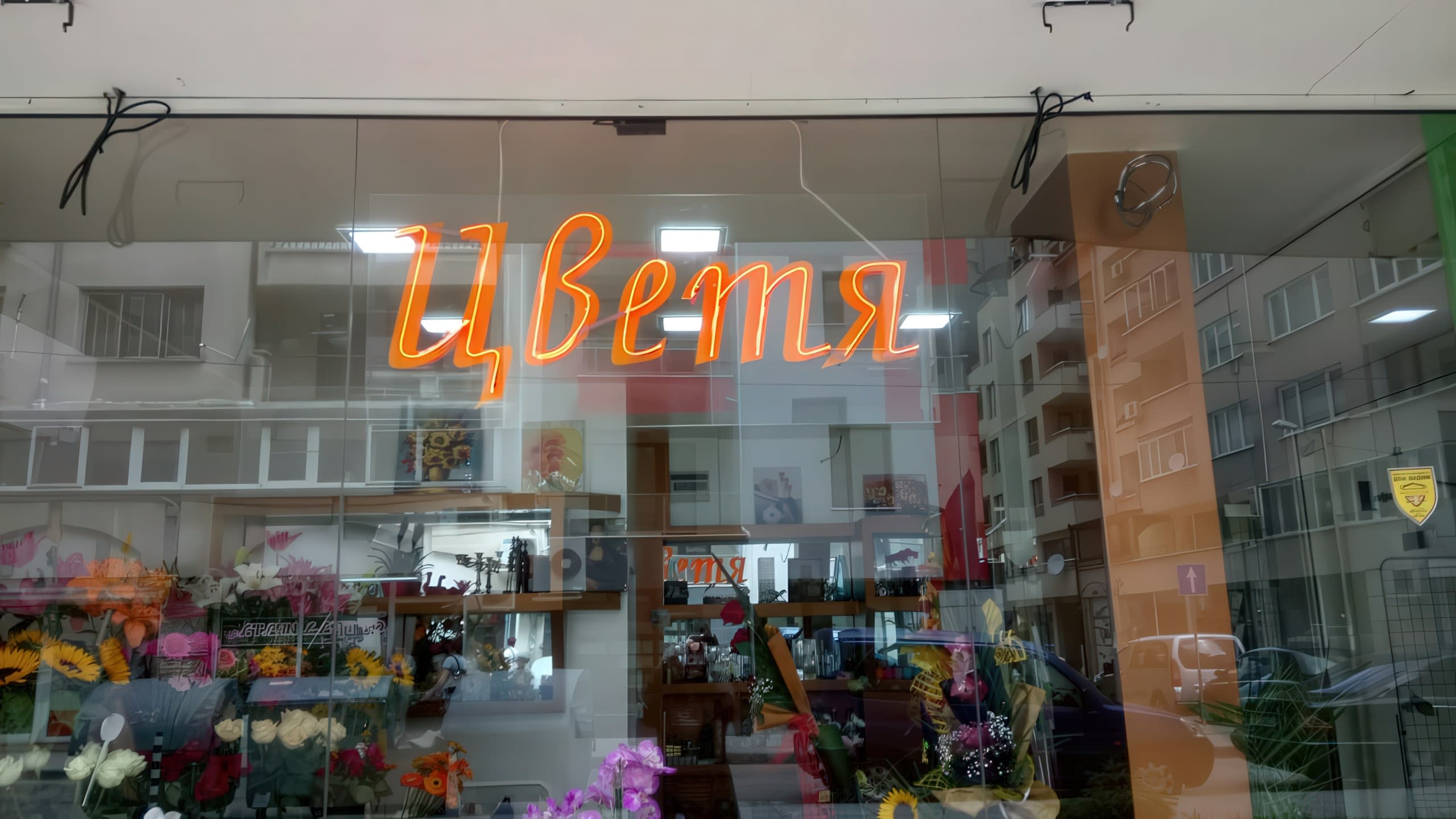 Неонов надпис "Цветя" в оранжев цвят, окачен на витрината на магазин за цветя през деня.