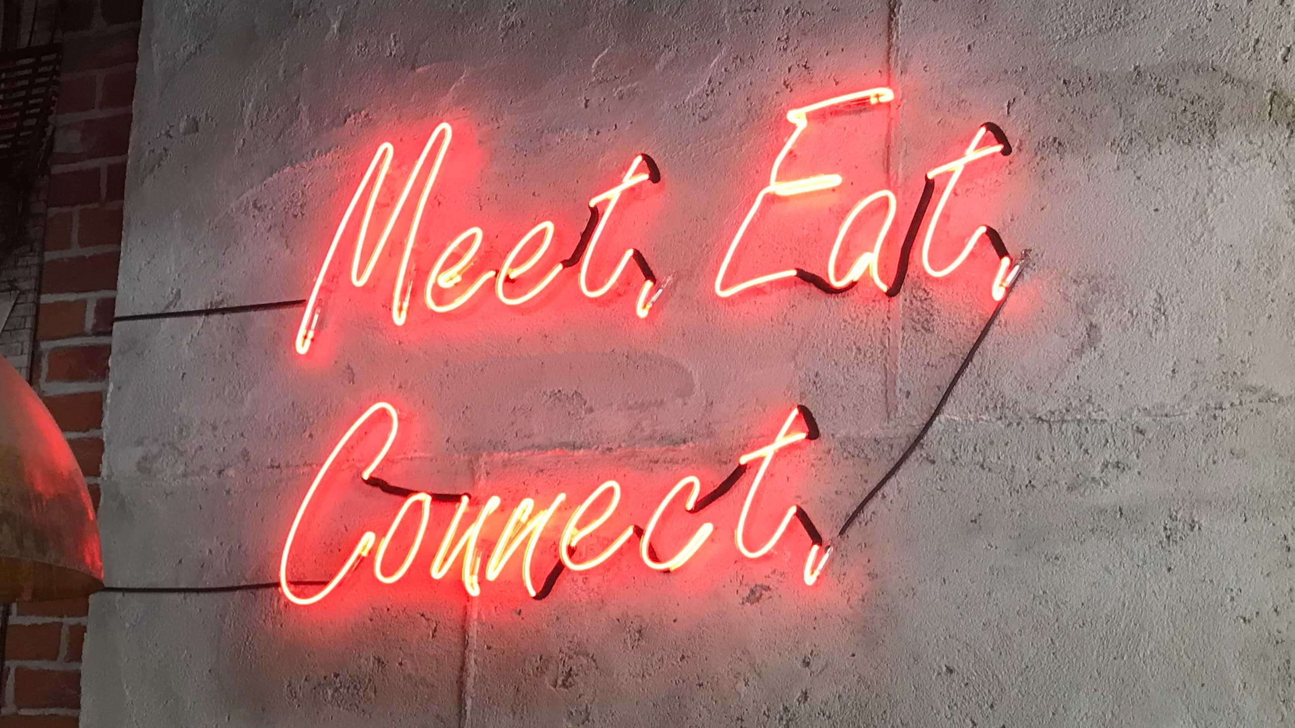 Интериорна неонова табела с ръкописен надпис "Meet, Eat, Connect", светеща в червено на бетонна стена.