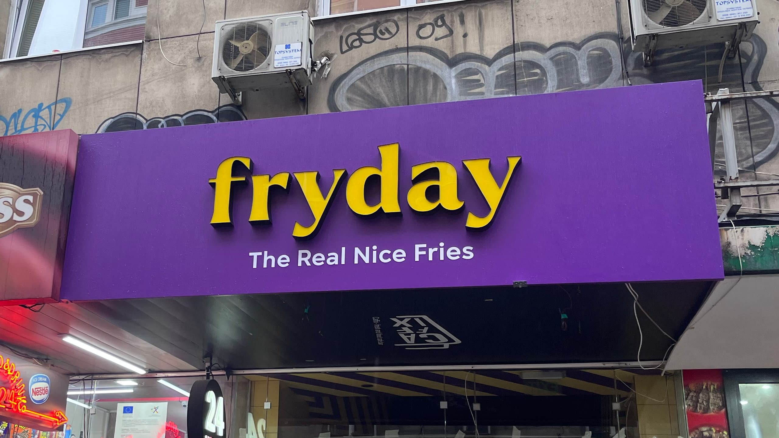 Табела на заведение за бързо хранене "fryday" с жълти обемни букви от плексиглас върху лилава основа.