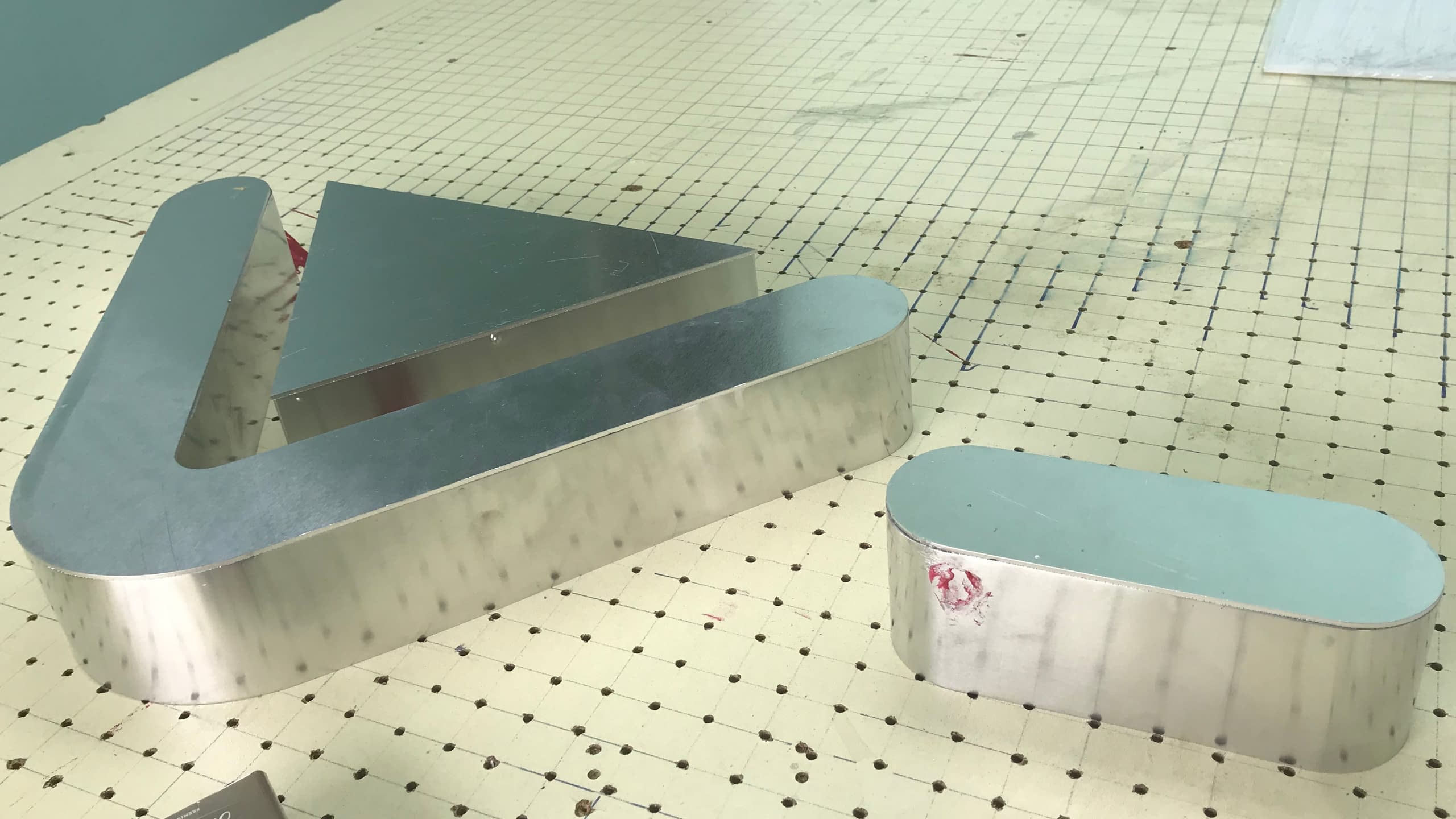 Машинно огънати алуминиеви бордове за обемни букви, оформени като буква V и буква O, поставени върху разграфена маса.