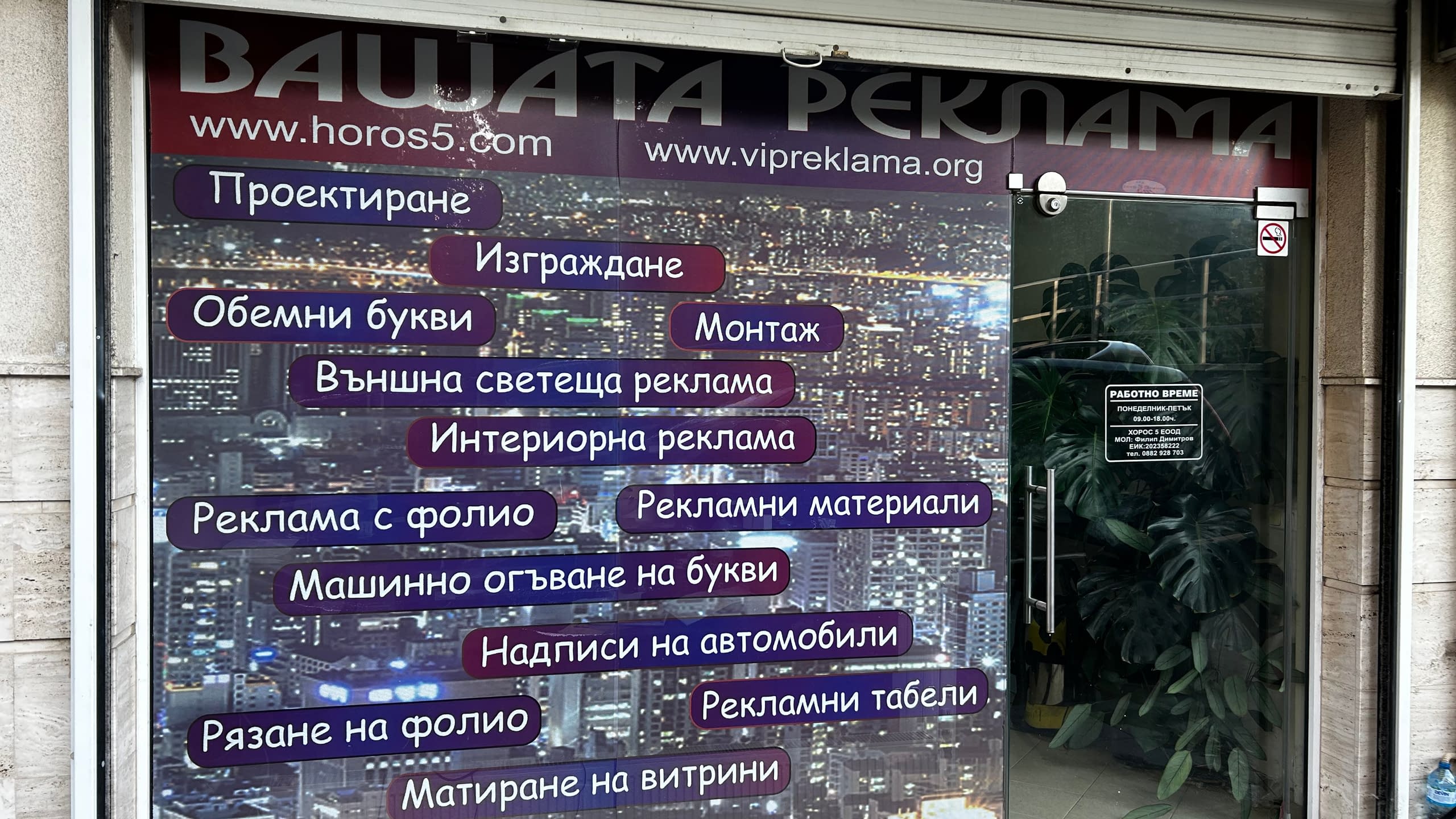 Витрина на рекламна агенция, брандирана с перфорирано фолио с нощен градски пейзаж и списък с предлагани услуги.