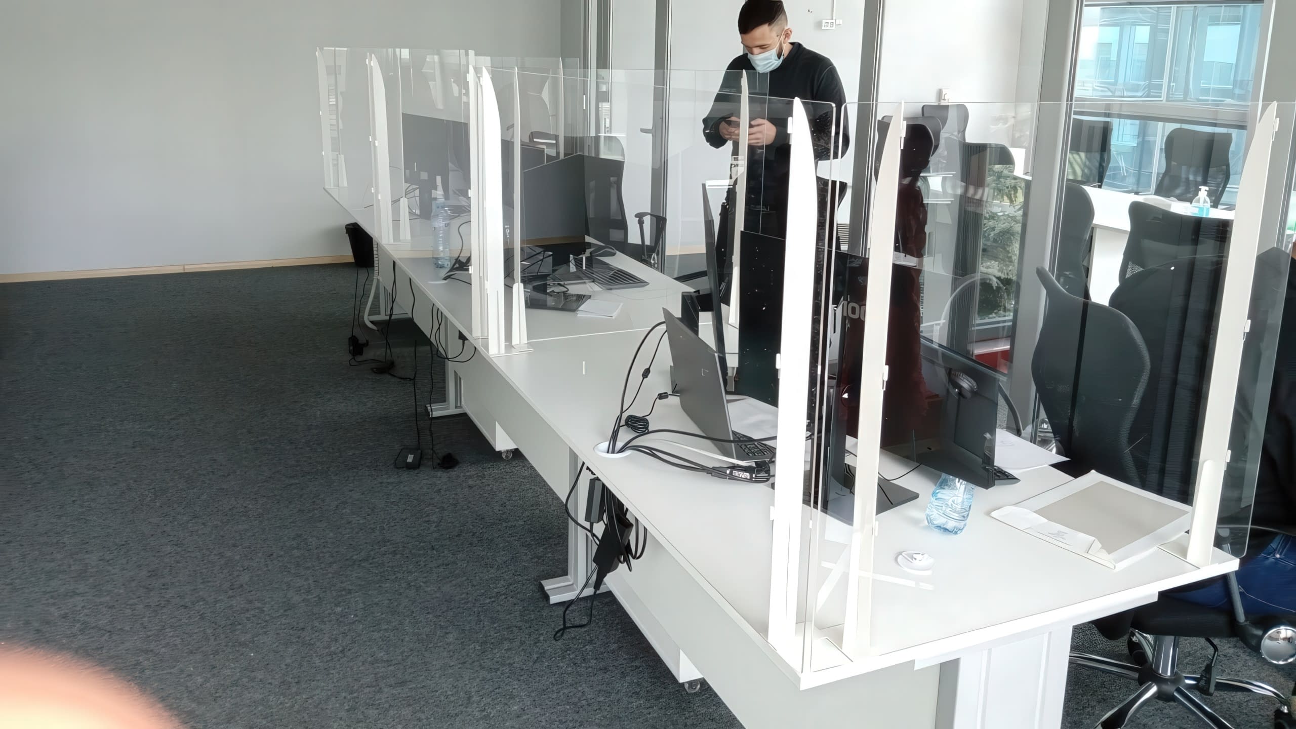 Модерен офис с бели бюра, разделени с прозрачни предпазни паравани от плексиглас.