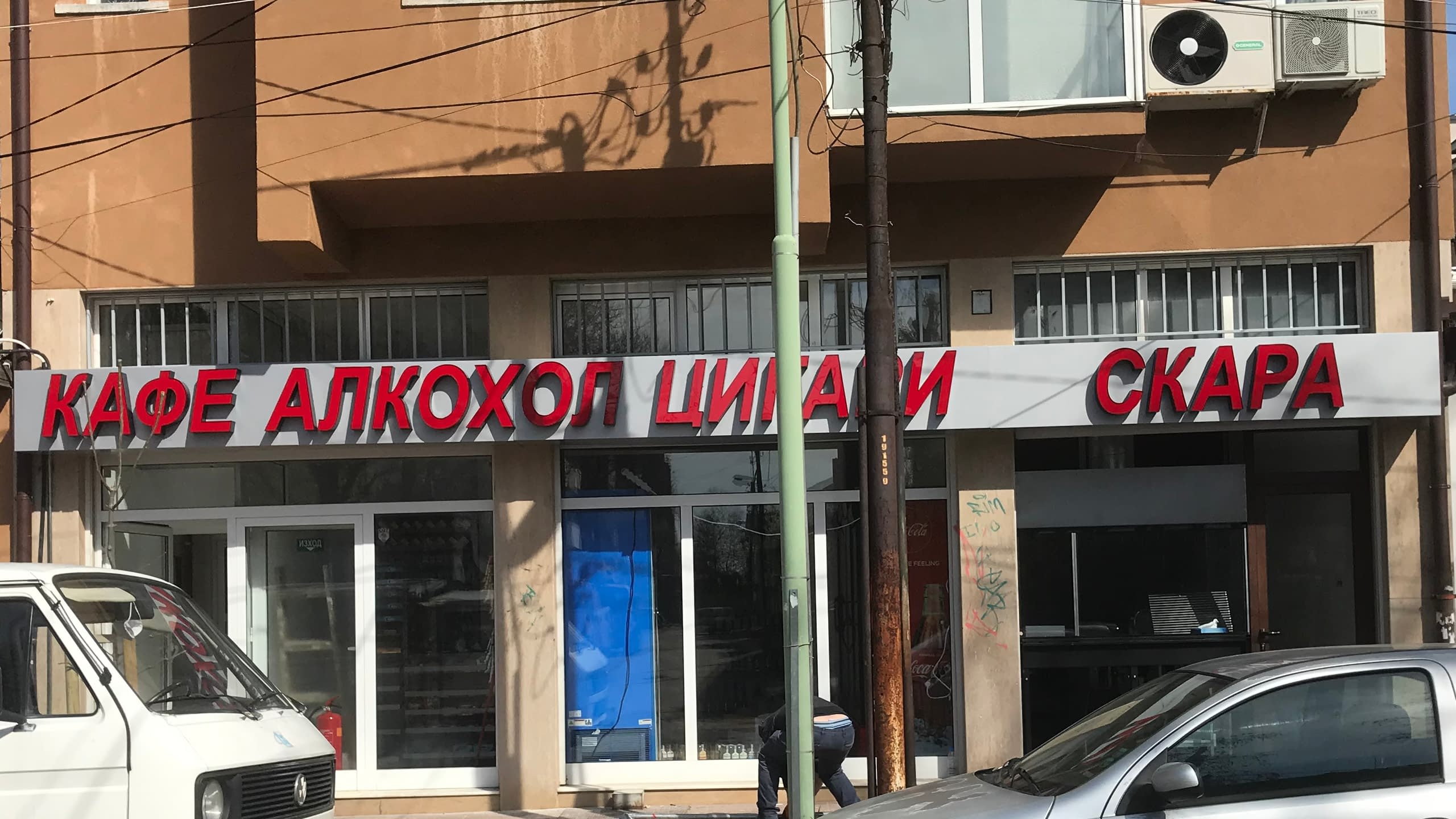 Дълга рекламна табела над магазин с червени обемни букви "Кафе Алкохол Цигари Скара".