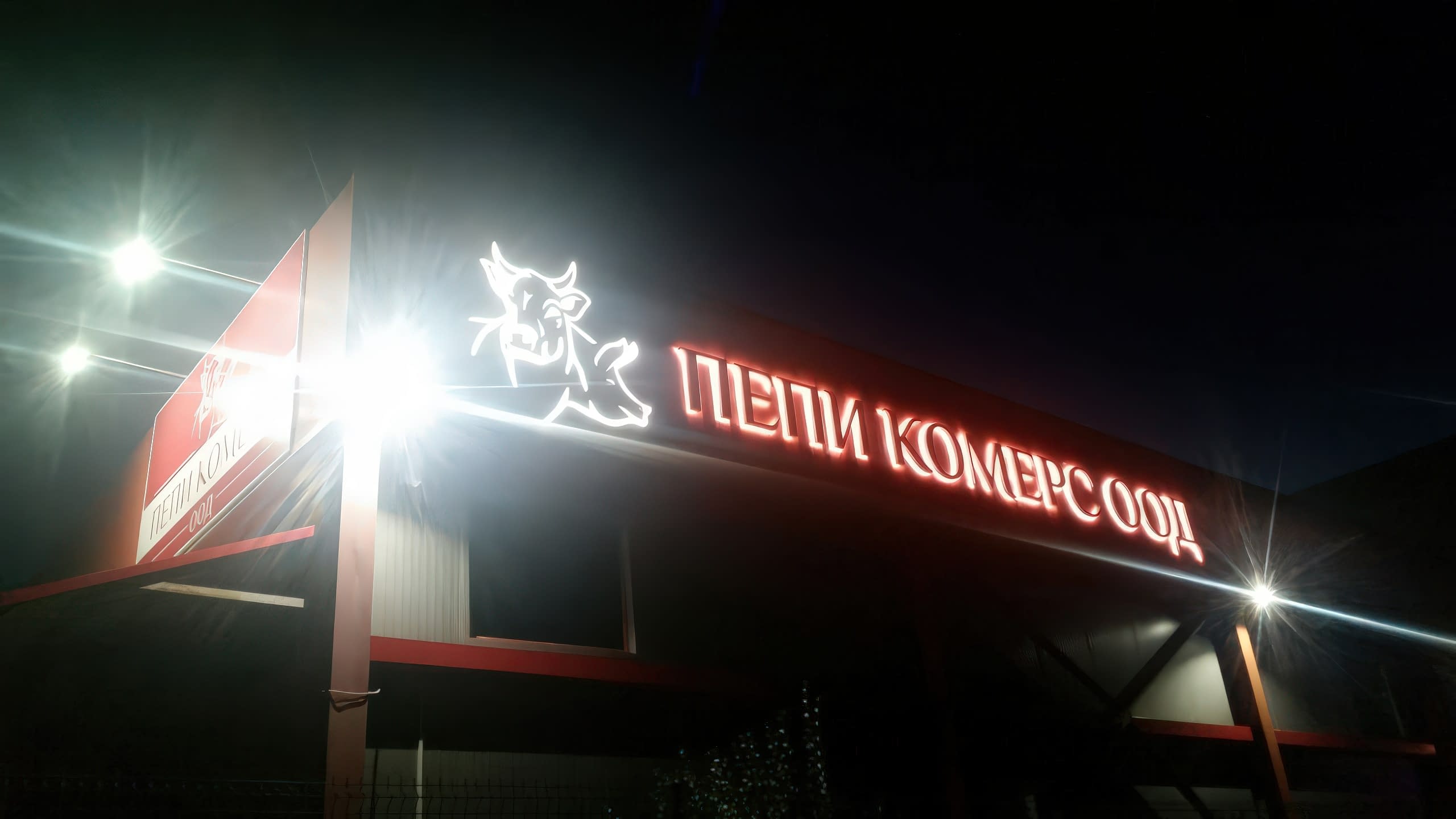 Нощна снимка на светеща покривна реклама с червени обемни букви "Пепи Комерс ООД" и светещо лого с крава.