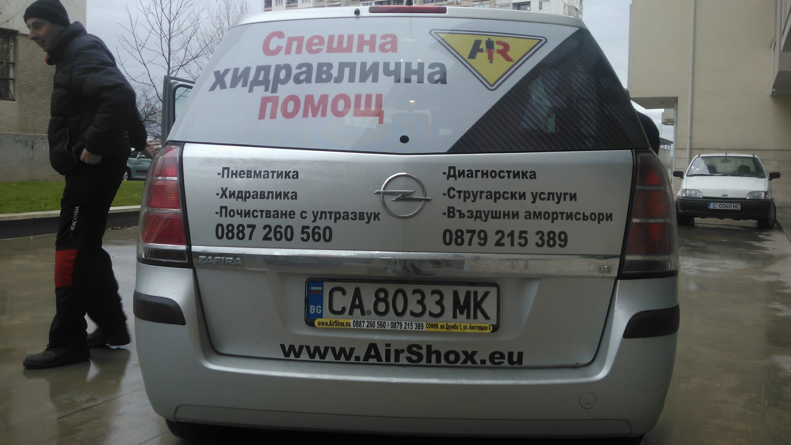 Задната част на сребърен Опел с рекламен надпис "Спешна хидравлична помощ" на стъклото и изписани телефонни номера.