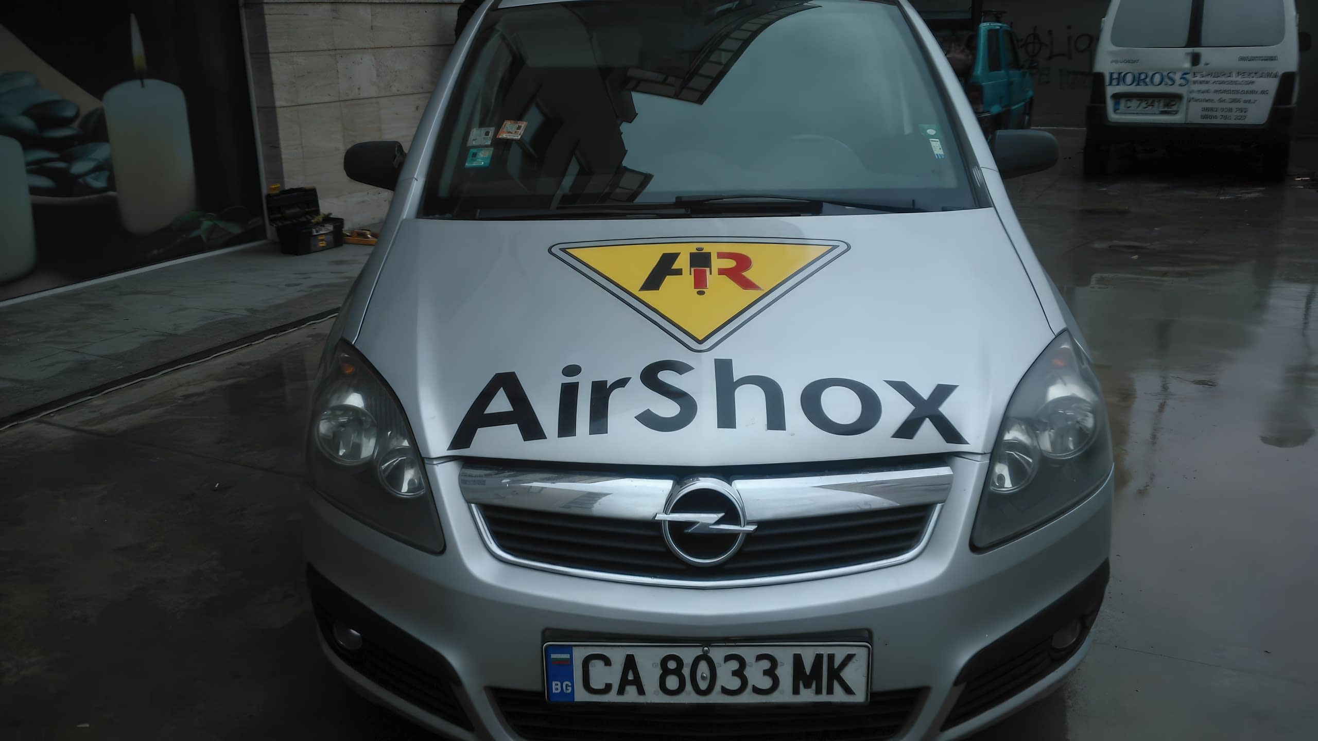 Преден план на капака на сребриста кола, върху който са поставени логото и името на фирма "AirShox".
