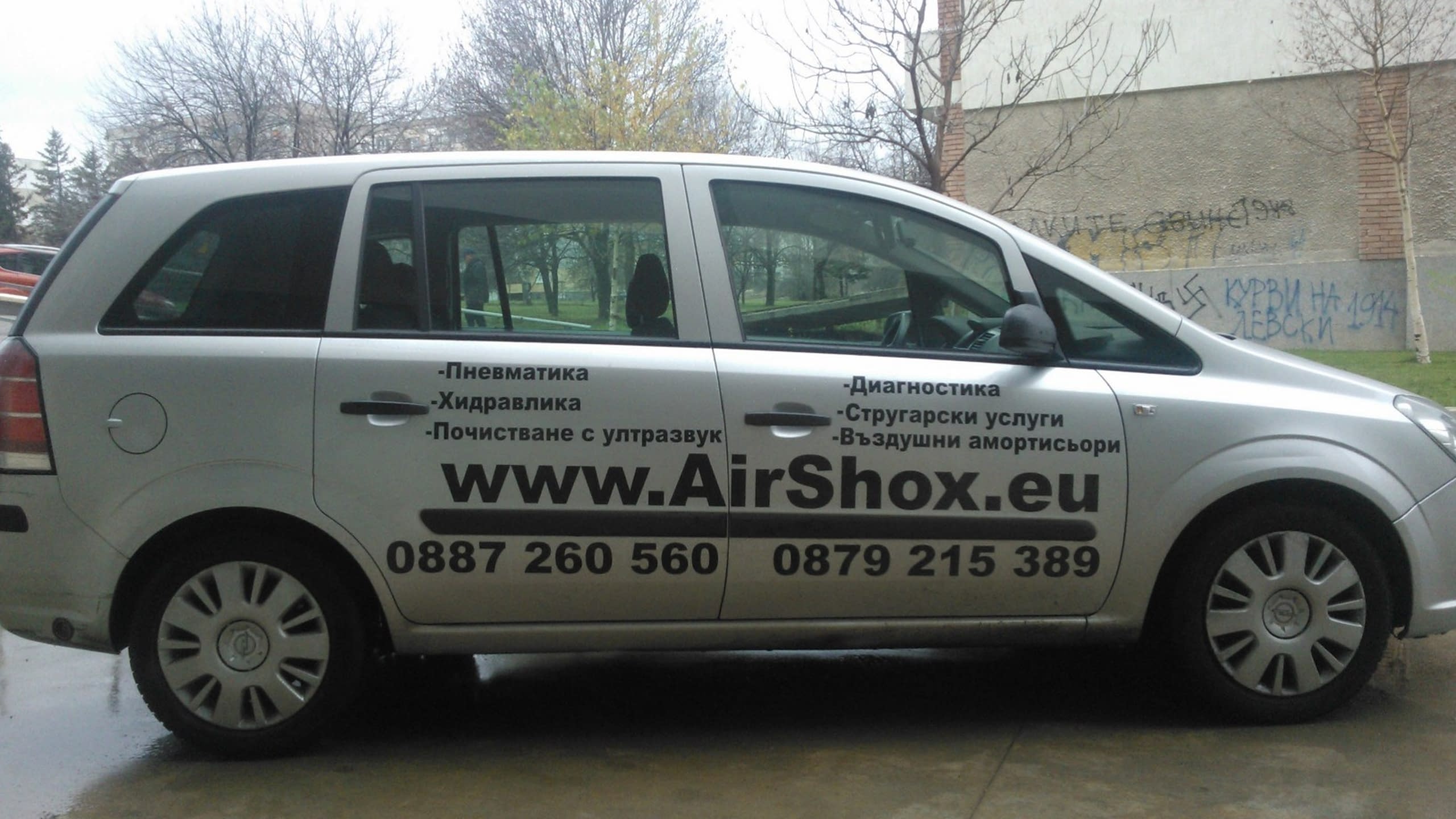 Страничен изглед на сребрист автомобил Опел Зафира с рекламни надписи по вратите, паркиран на открито.