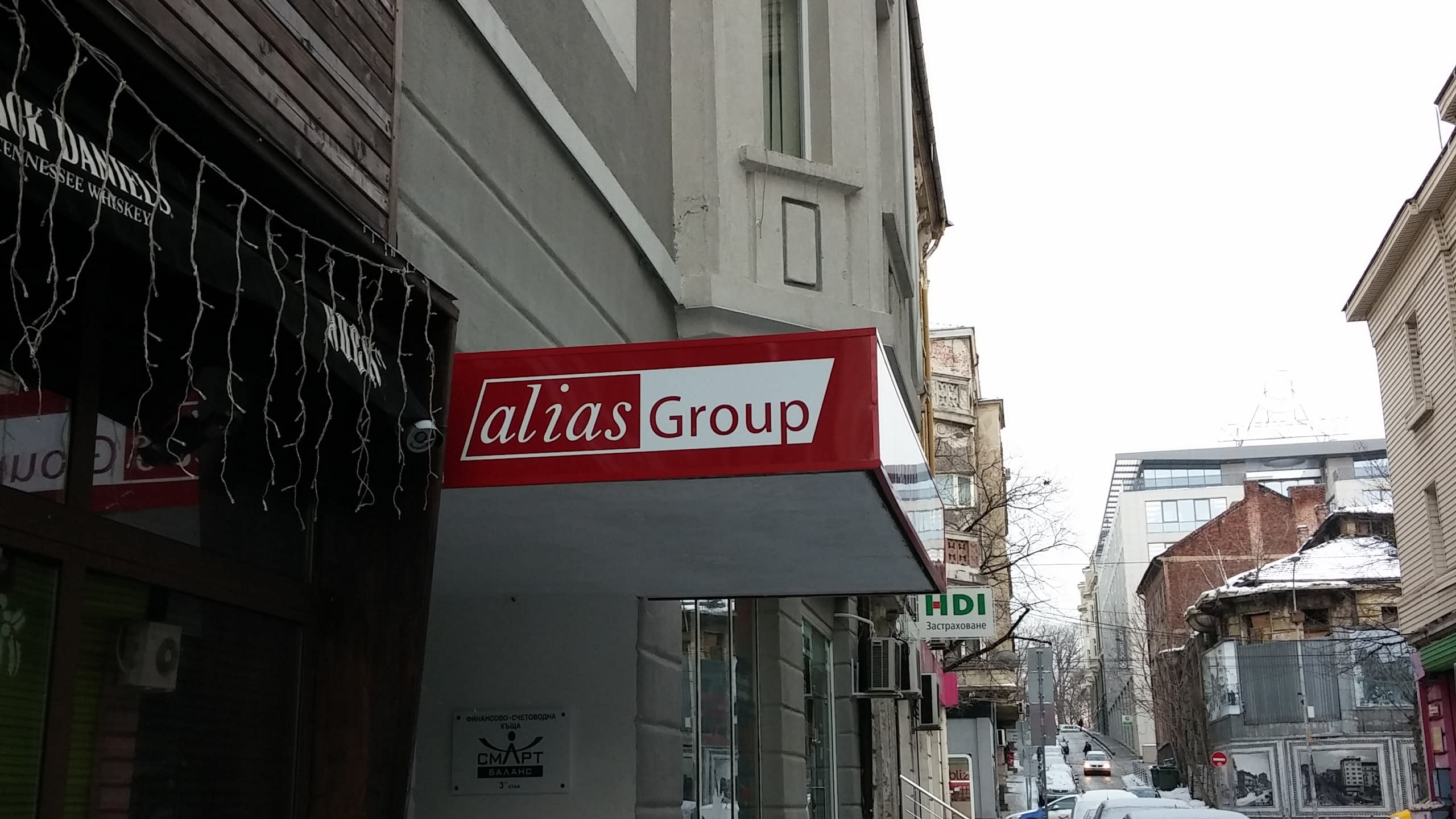 Страничен изглед на червена фирмена табела, която изпъква от фасадата на сграда на градска улица.