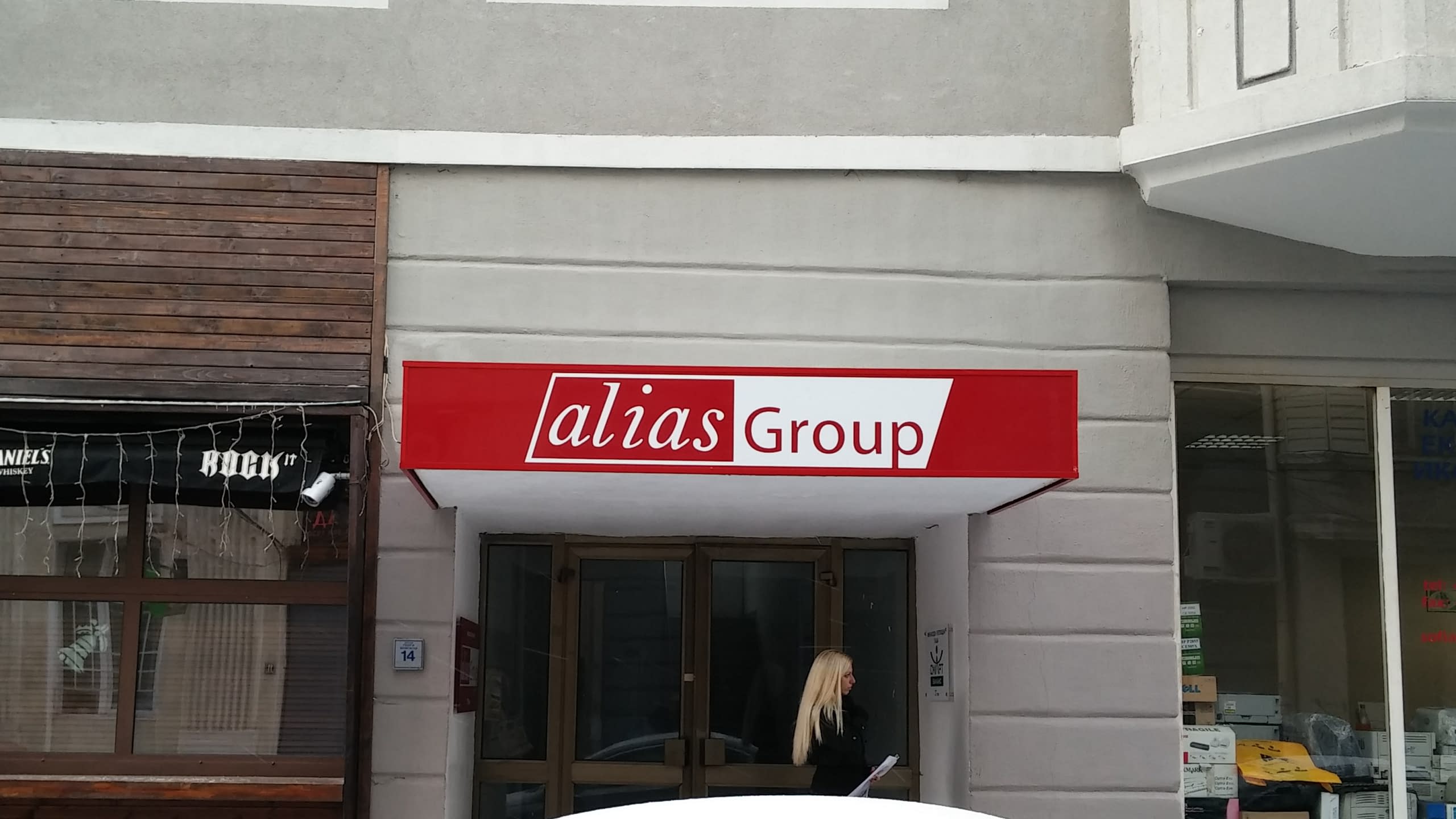 Фронтален изглед на табела тип козирка с логото на "alias Group" над входа на офис.