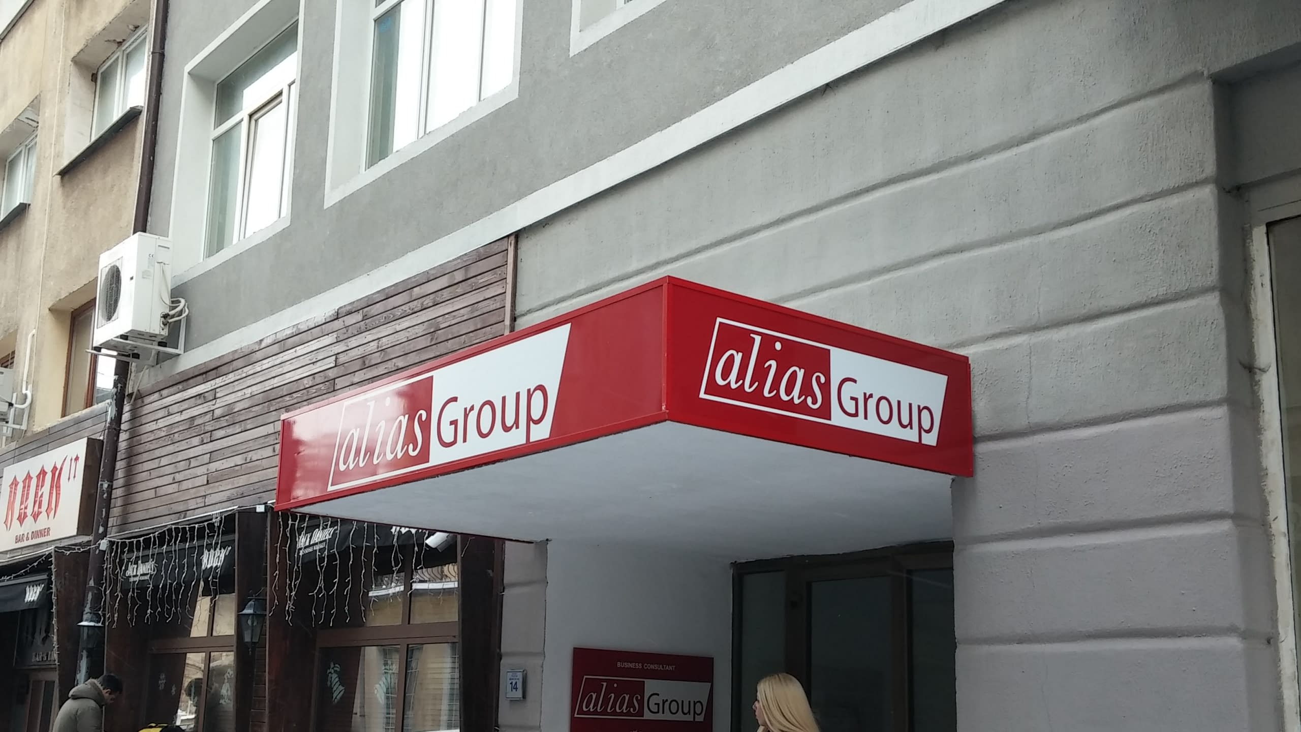 Обемна табела в червено и бяло с фирмено лого, снимана отдолу под ъгъл, за да се видят трите ѝ страни.