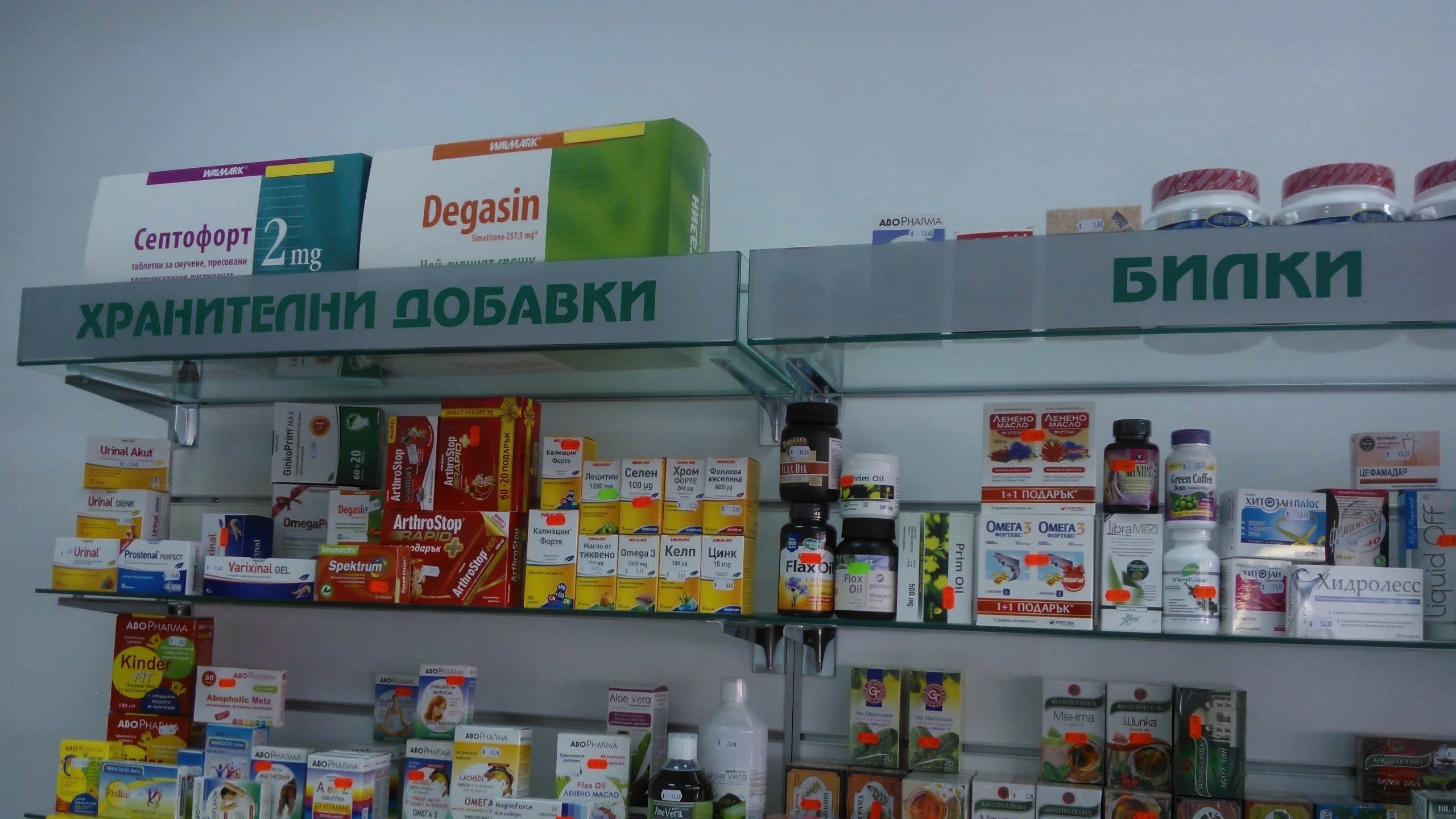 Близък план на рафтове с лекарства и хранителни добавки, надписани със зелени букви "Хранителни добавки" и "Билки".
