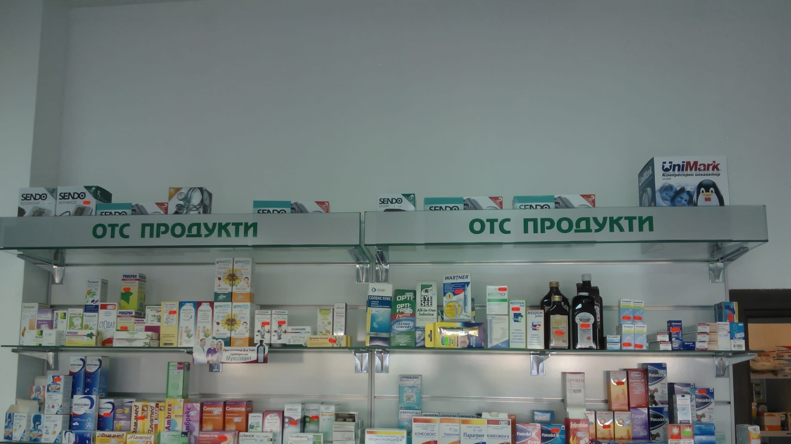 Две съседни секции с рафтове в аптека, обозначени с надпис "ОТС Продукти".