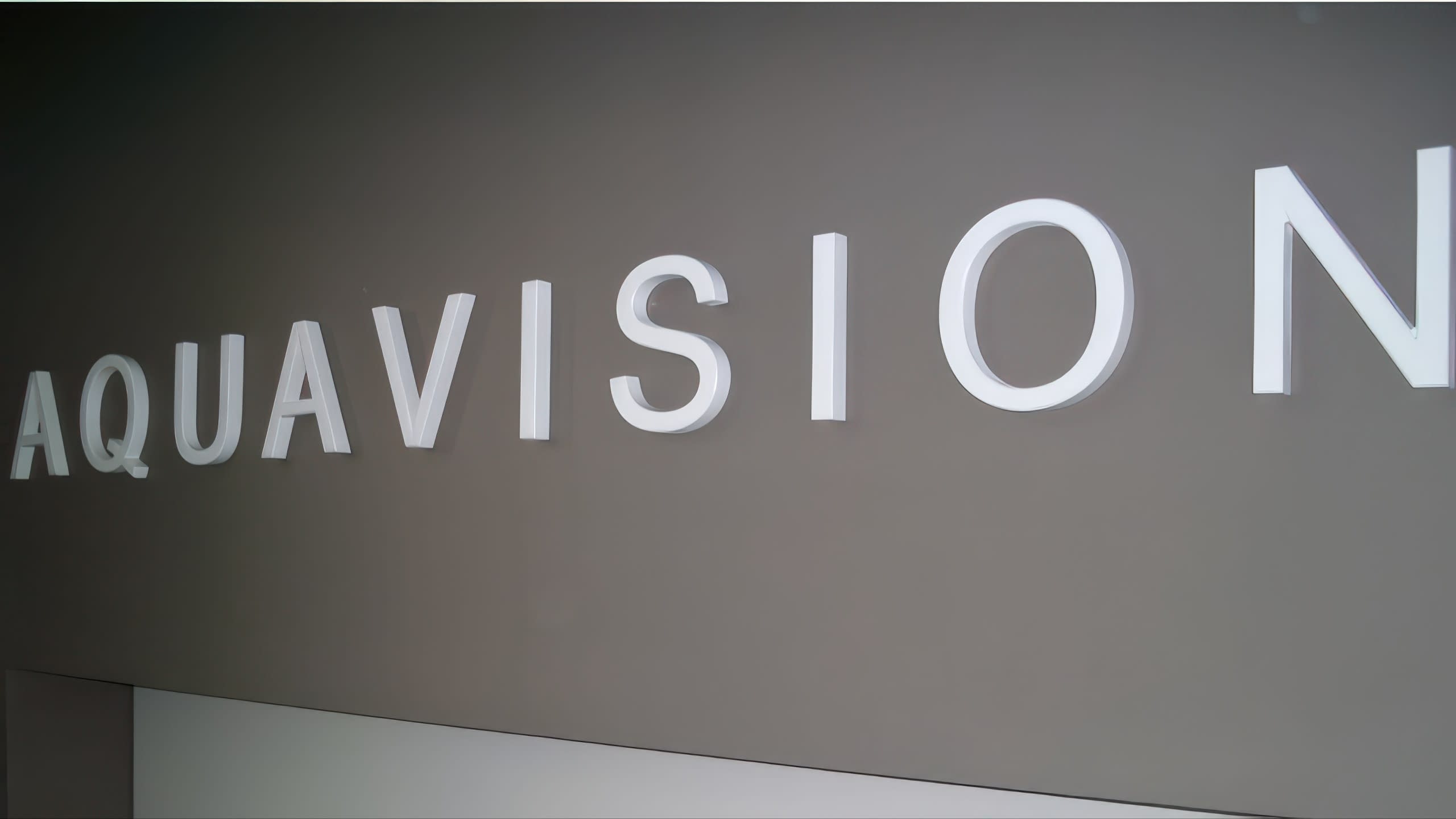 Бели букви с надпис AQUAVISION, снимани под ъгъл, за да се види тяхната дебелина и монтаж на стената.
