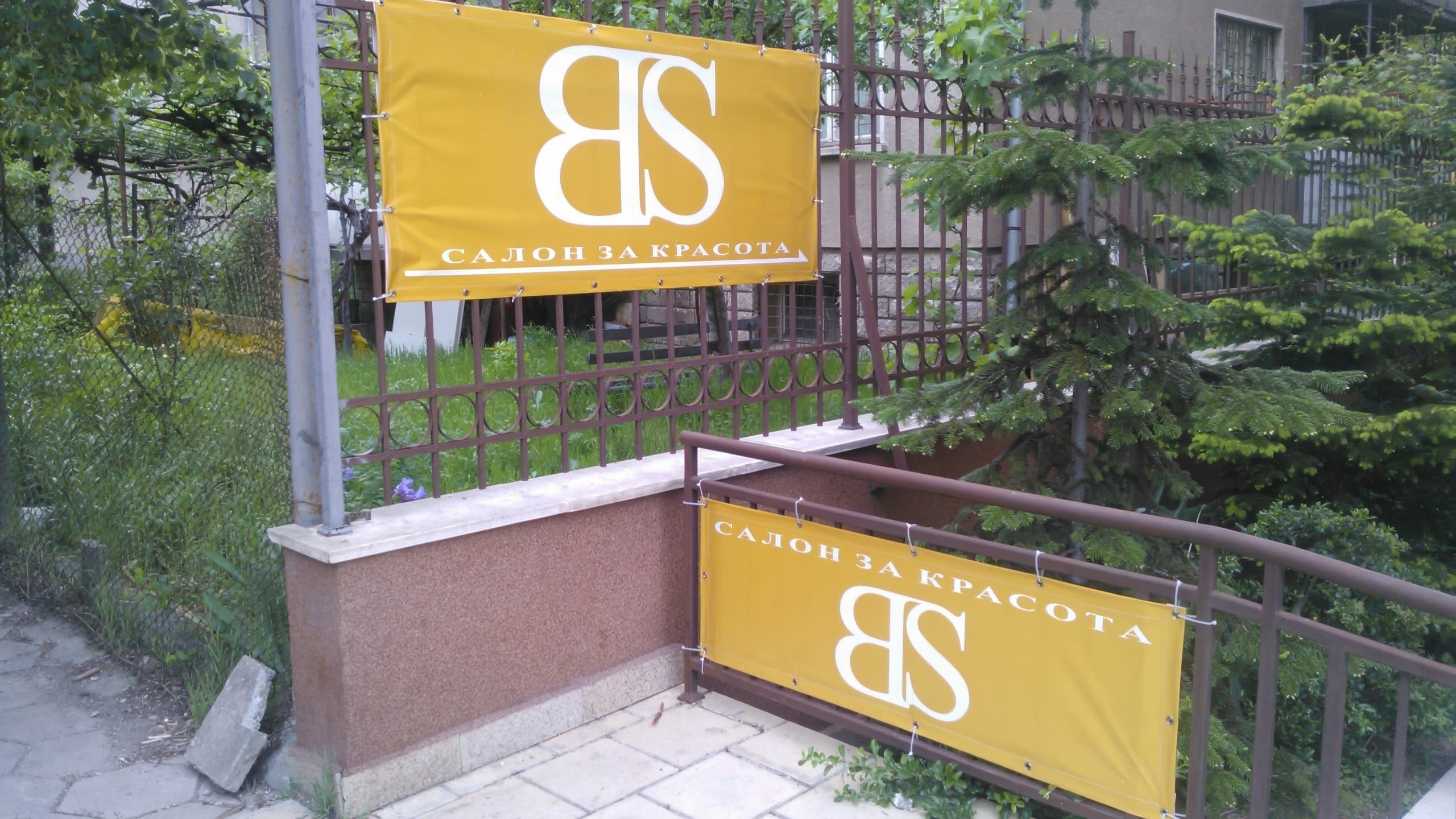 Два жълти транспаранта за салон за красота, закачени на ограда пред къща със зеленина.