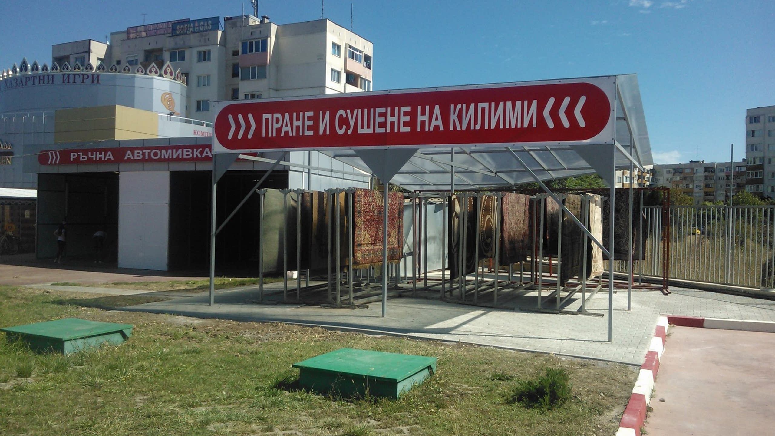Червен винилов транспарант с надпис "Пране и сушене на килими", монтиран на метална конструкция на открито.