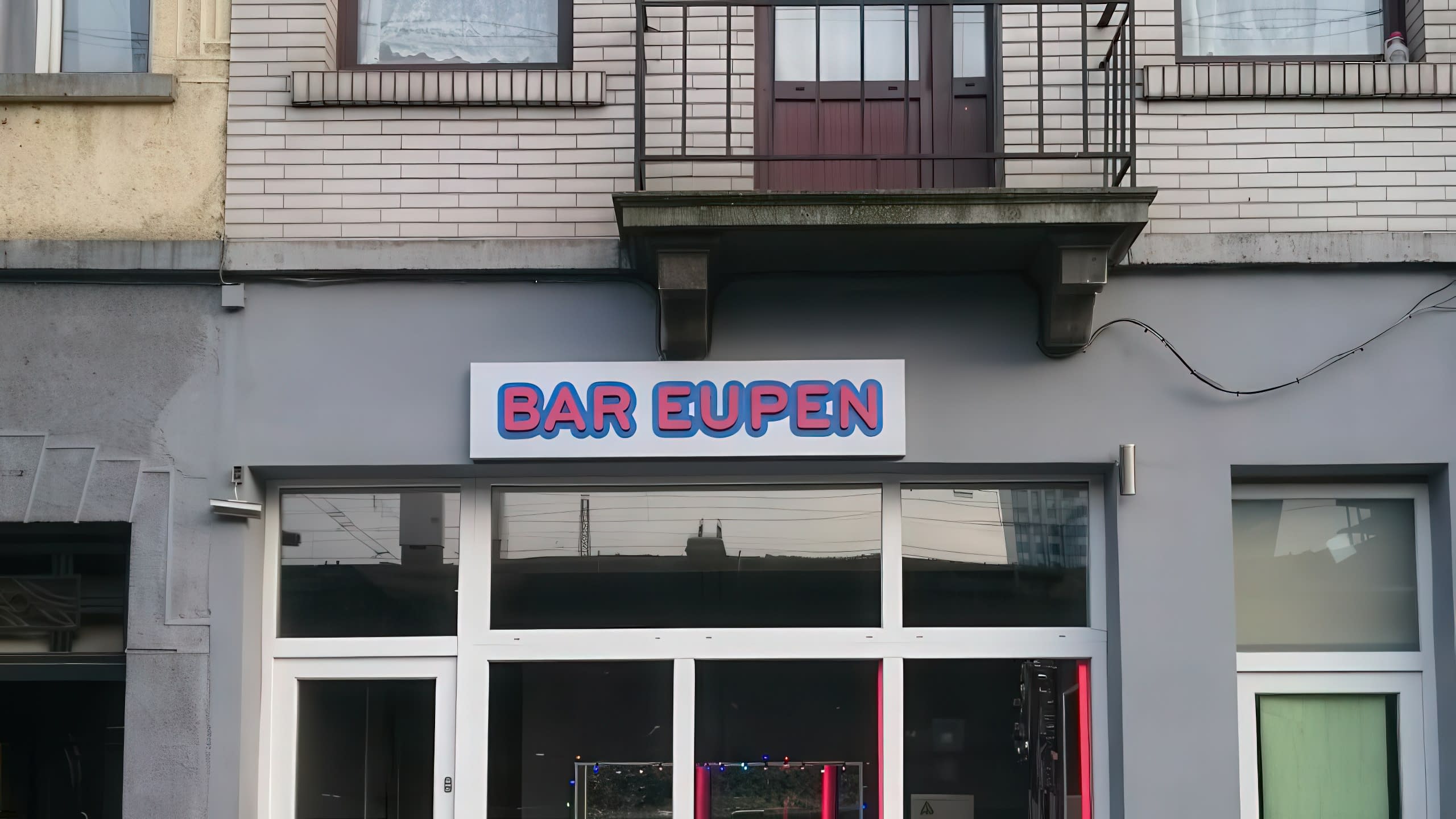 Нощна снимка на входа на „Bar Eupen“, над който е поставена осветената табела с името му.
