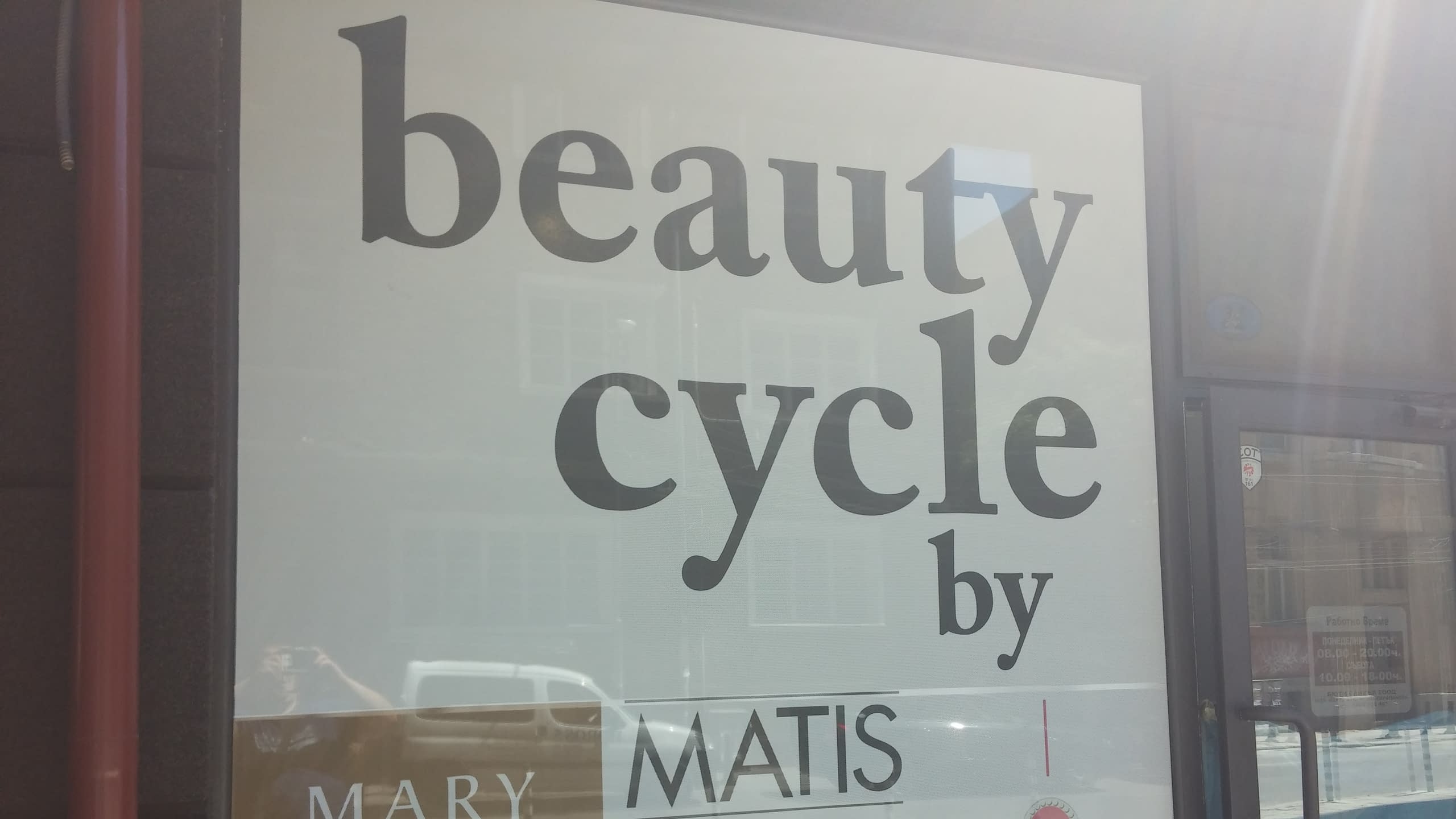 Близък план на надписа „beauty cycle“ върху фолио за витрина, с отражения на сгради и превозно средство.