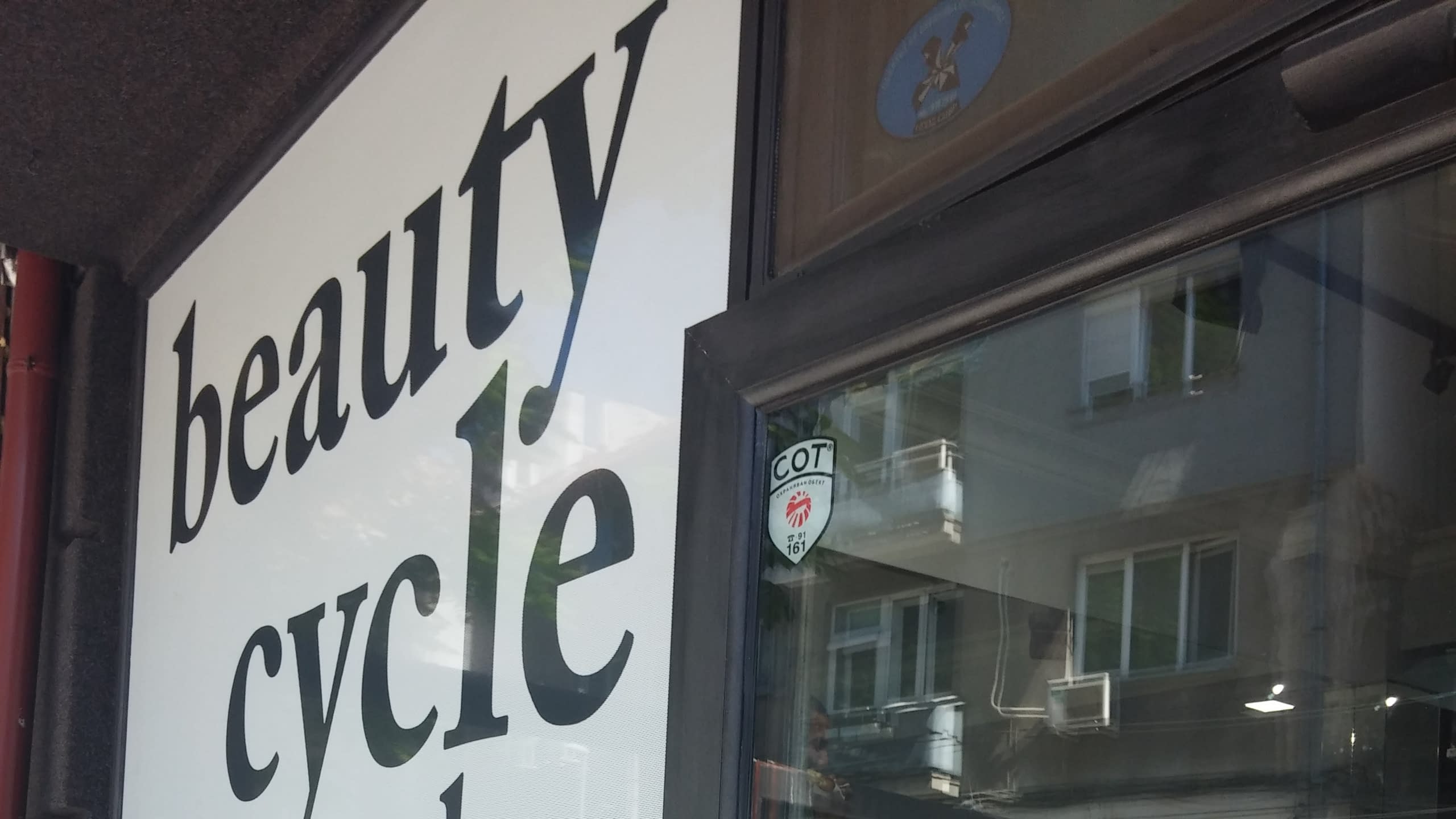 Ъглов изглед към входа на салон, показващ част от брандираната витрина и отражение в стъклената врата.