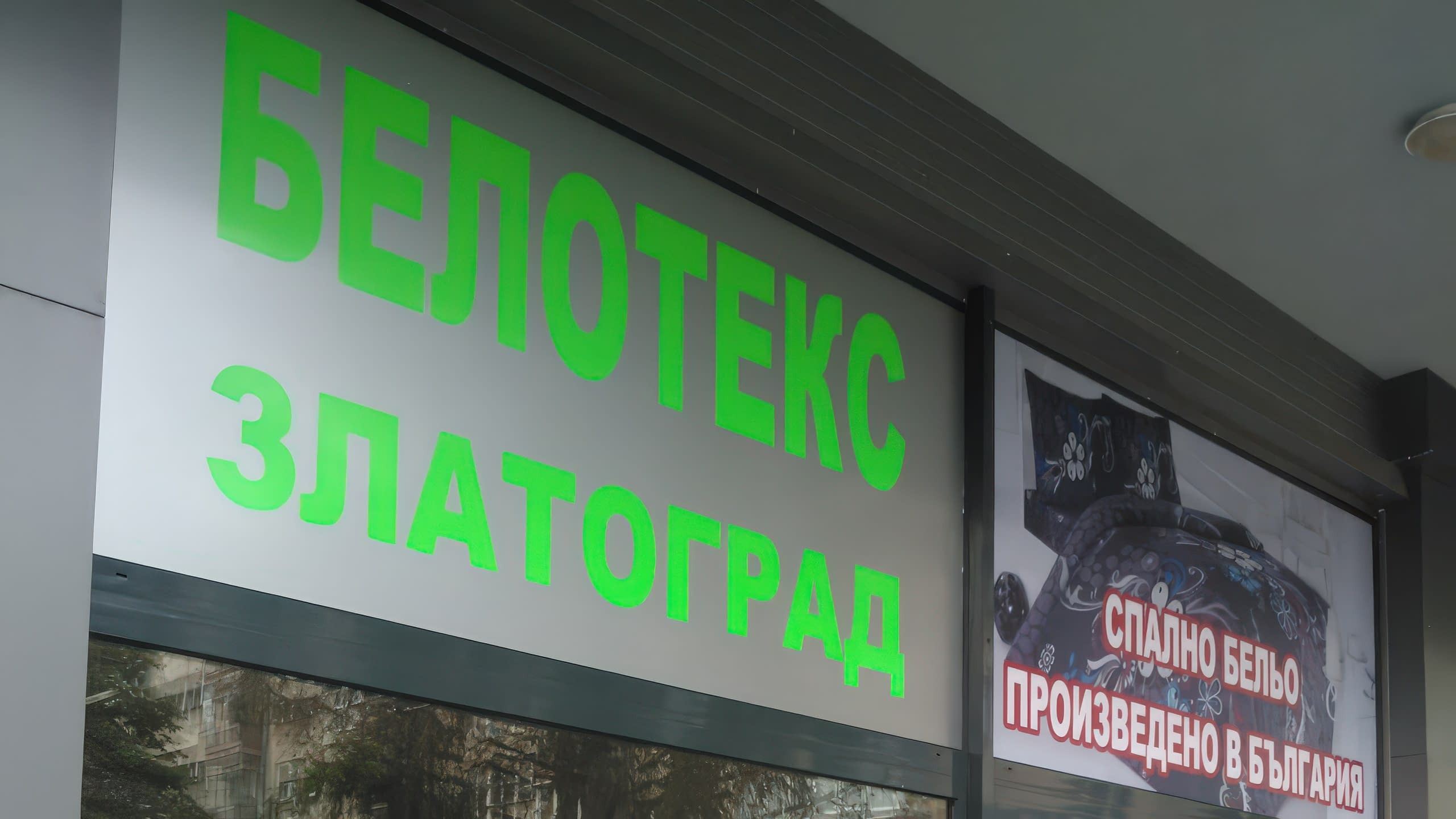 Два панела с фолио на витрина, погледнати под ъгъл – единият със зелен надпис, другият със снимка на спално бельо.