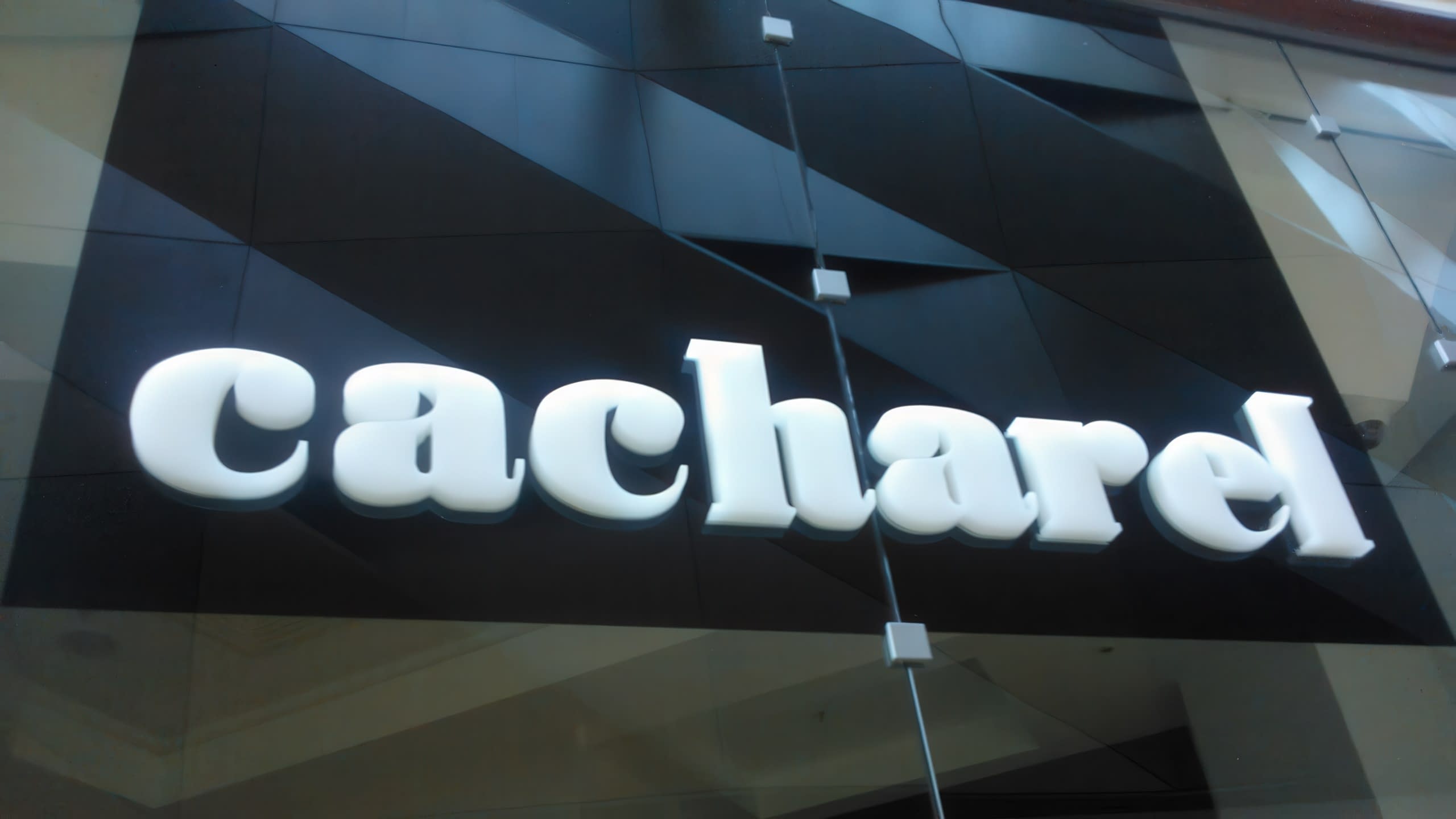 Надпис „cacharel“ с бели обемни букви, разположен върху черна повърхност с геометрични форми.