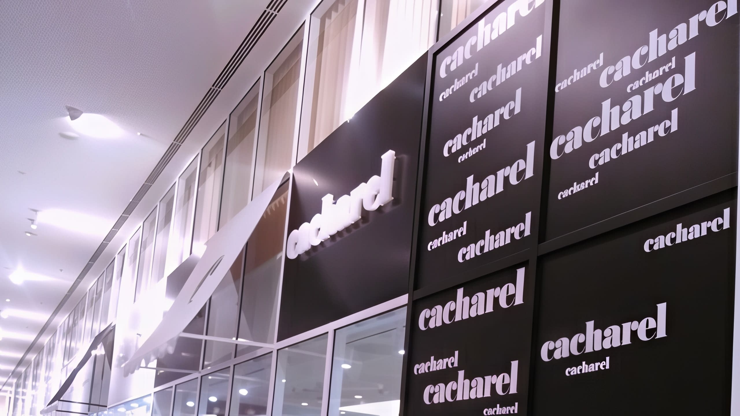 Изглед по дължината на витрина на магазин, брандирана с основна табела и панели с повтарящо се лого на Cacharel.