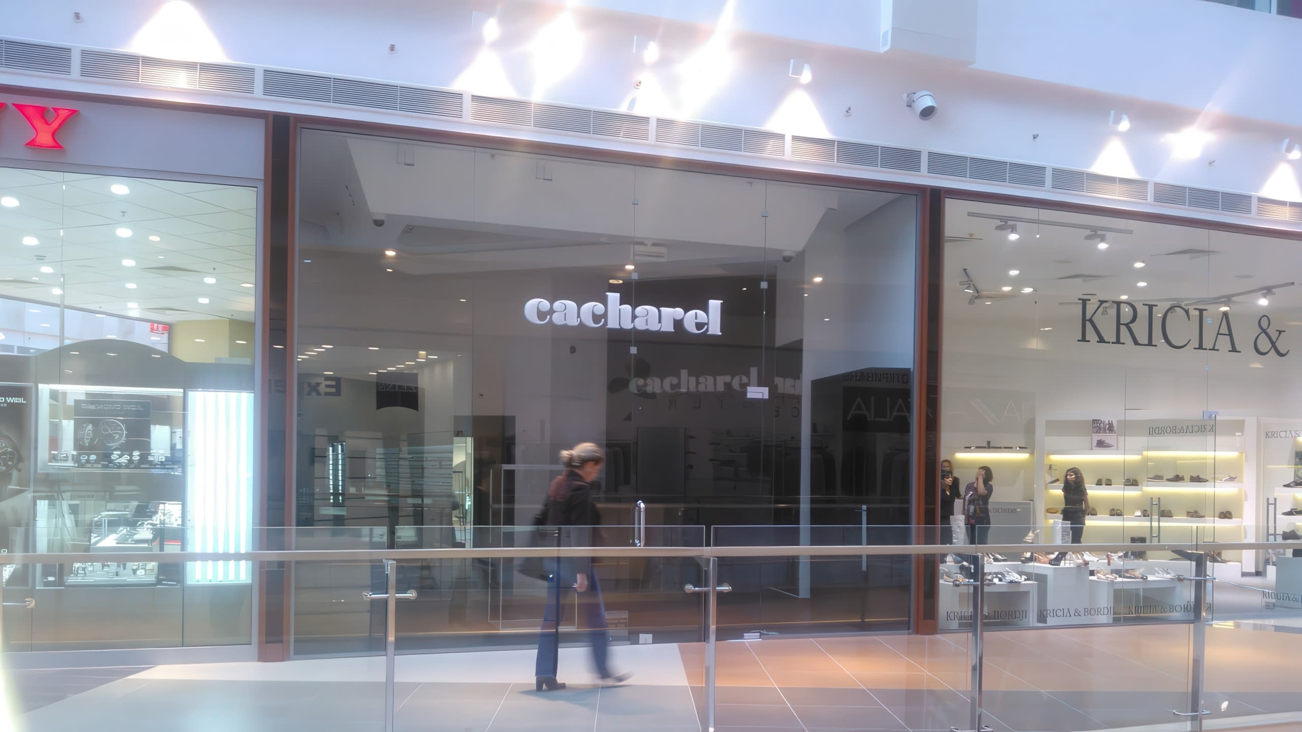 Изглед към витрините на магазини в търговски център, включително Cacharel, с човек, който преминава по коридора.