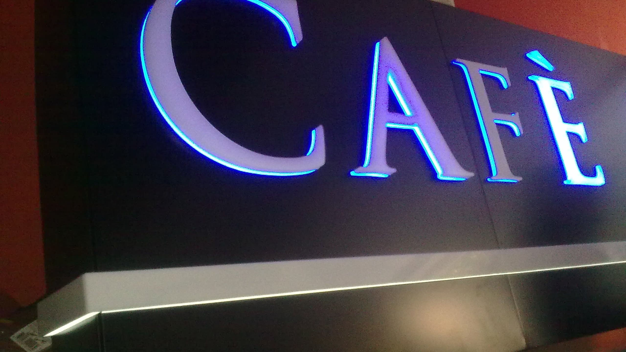 Надпис „CAFÈ“ с бели букви, осветени със синя контурна светлина, върху тъмна табела.
