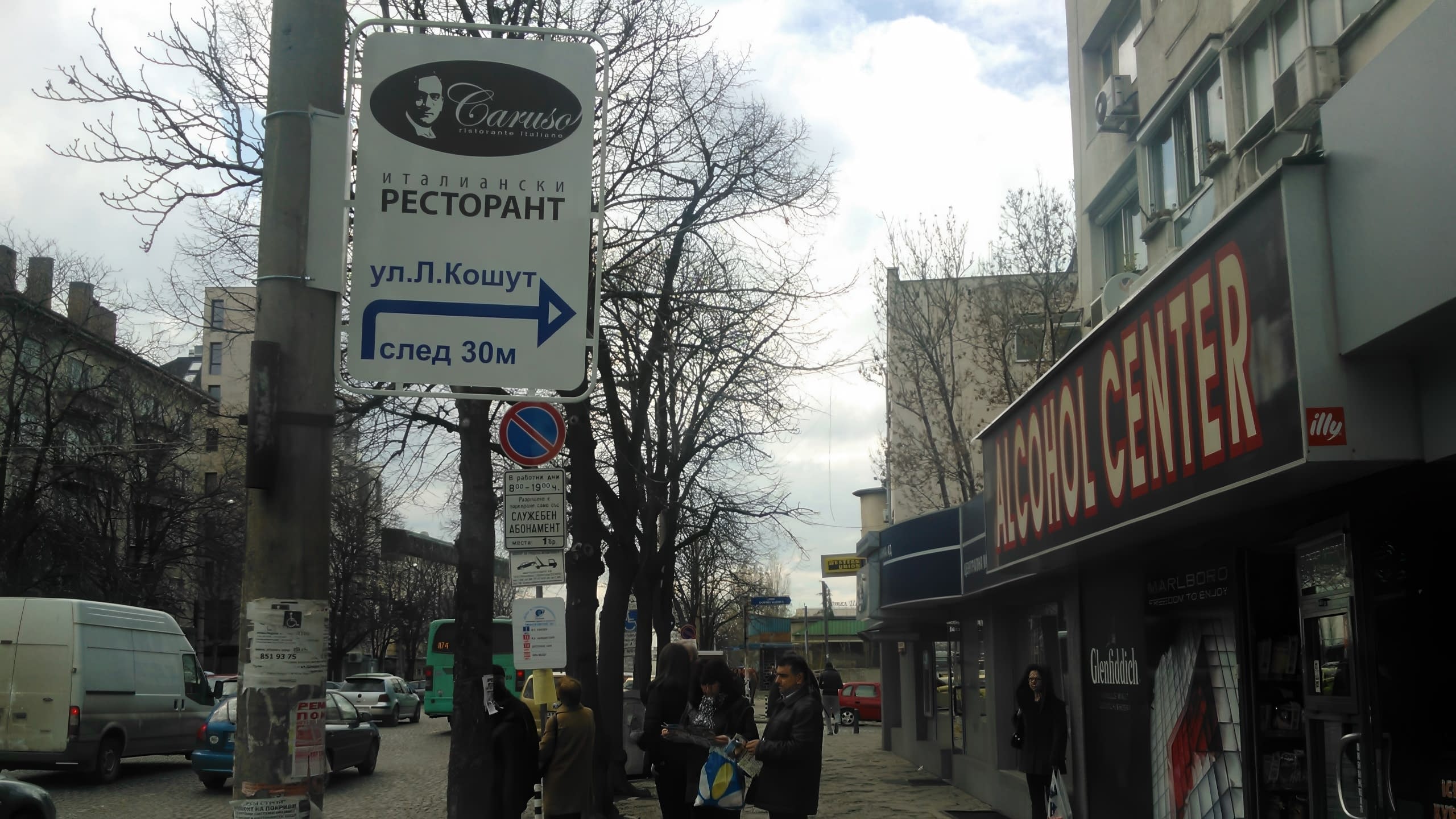 Табела на стълб до оживена улица с магазини, която насочва към ресторант Caruso на ул. „Л. Кошут“.