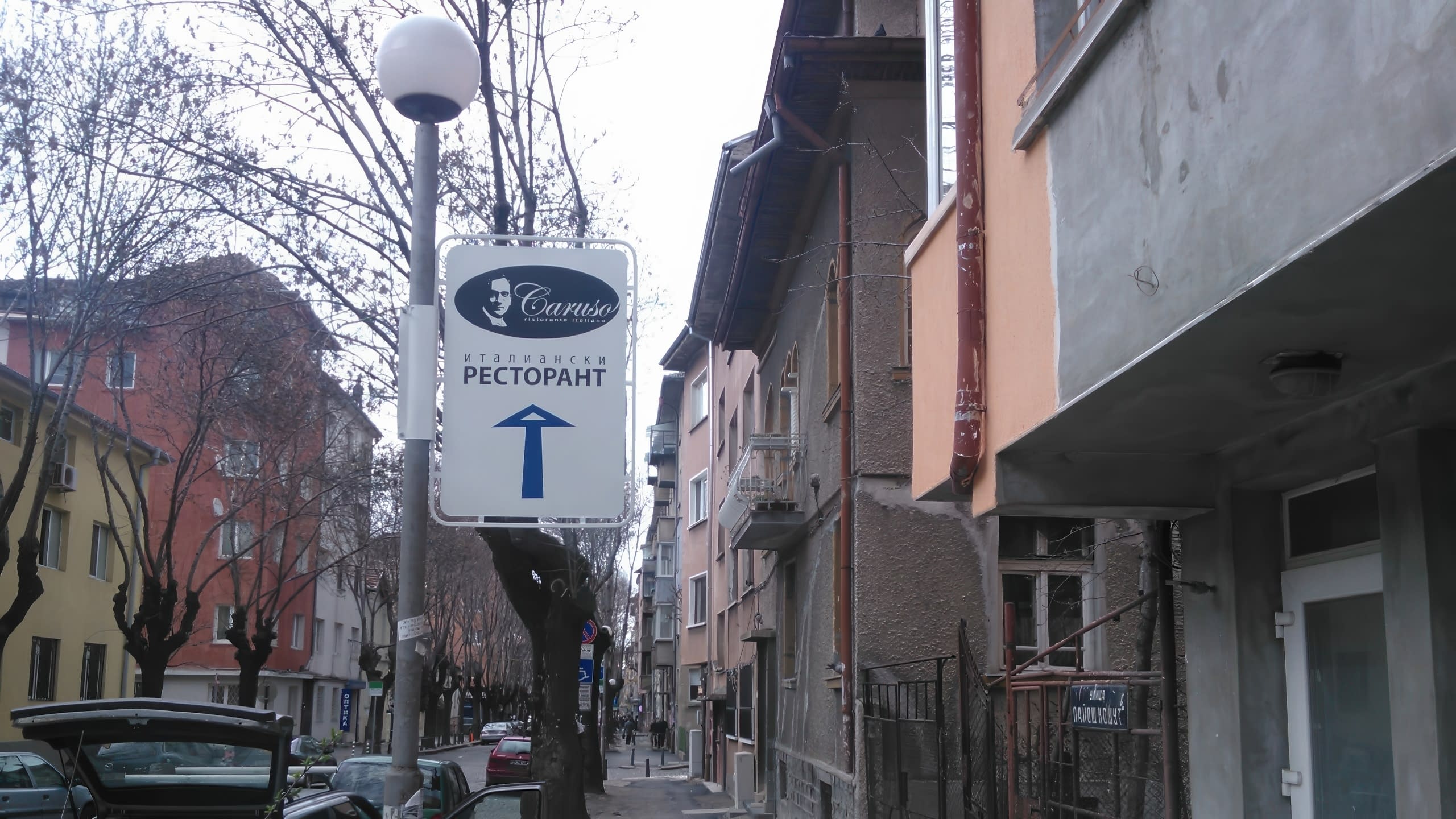 Улична табела на стълб с голяма синя стрелка, сочеща право напред по улица със стари сгради.