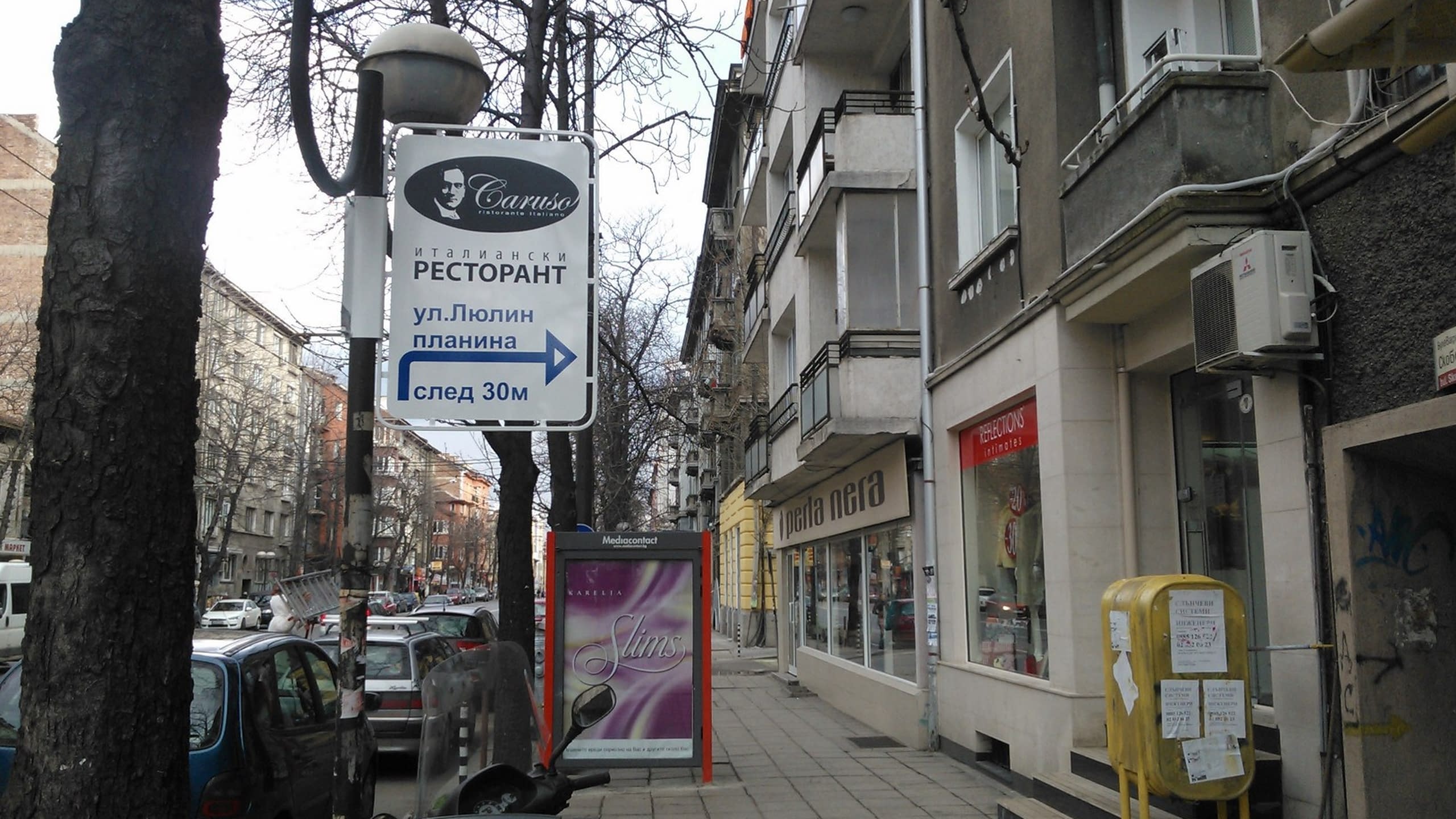 Бяла правоъгълна табела, монтирана на стълб на улица, с надписи и стрелка, указващи посоката към ресторант на ул. „Люлин планина“.