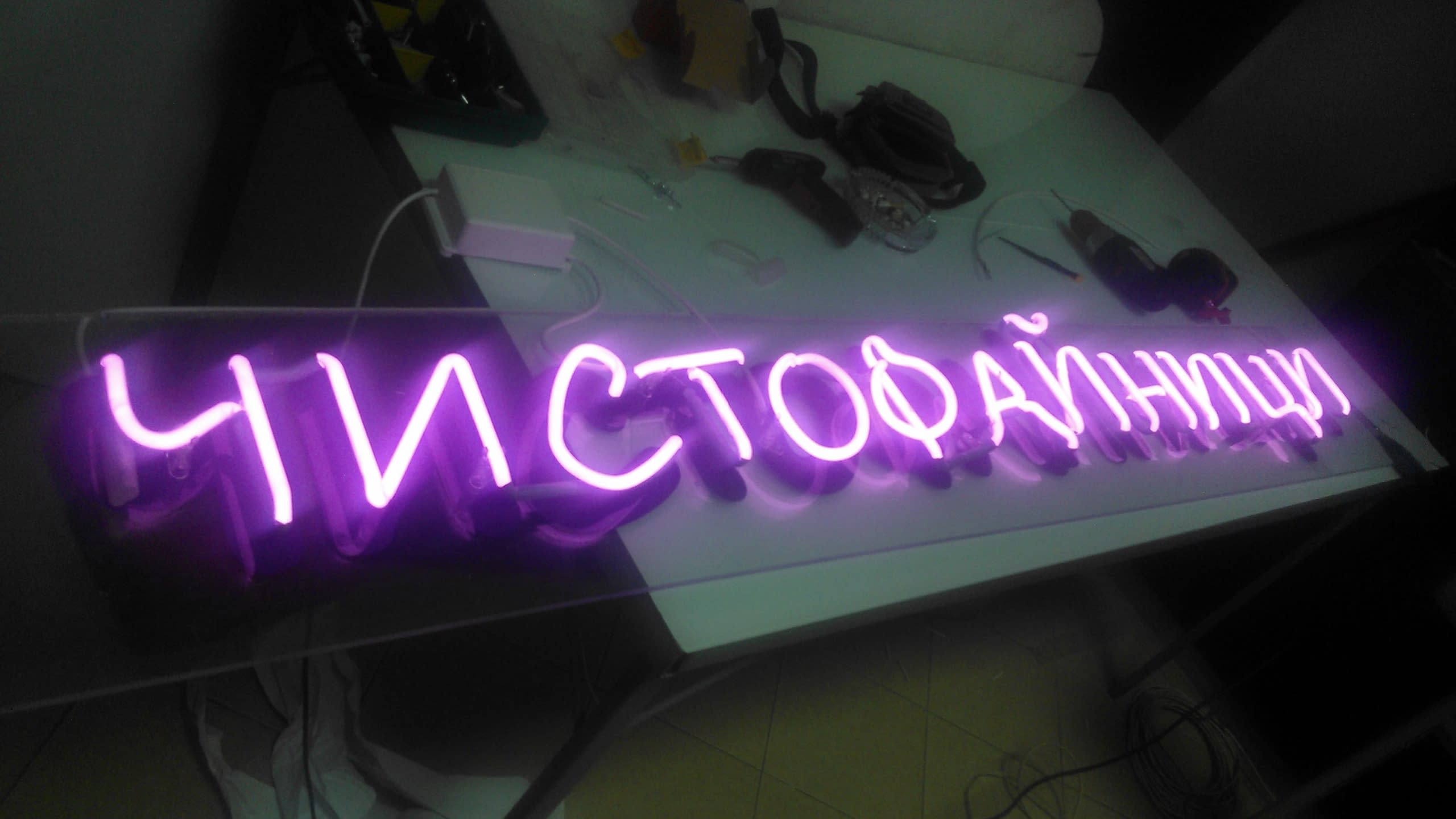 Лилав светещ надпис, поставен на работна маса в ателие, заобиколен от инструменти, захранващ блок и кабели.