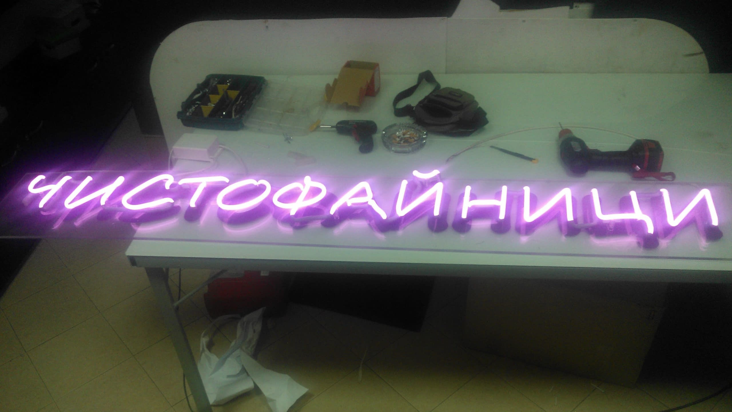 Светещ надпис в лилаво, разположен върху бяла маса в работилница, с различни инструменти като бормашини на заден план.