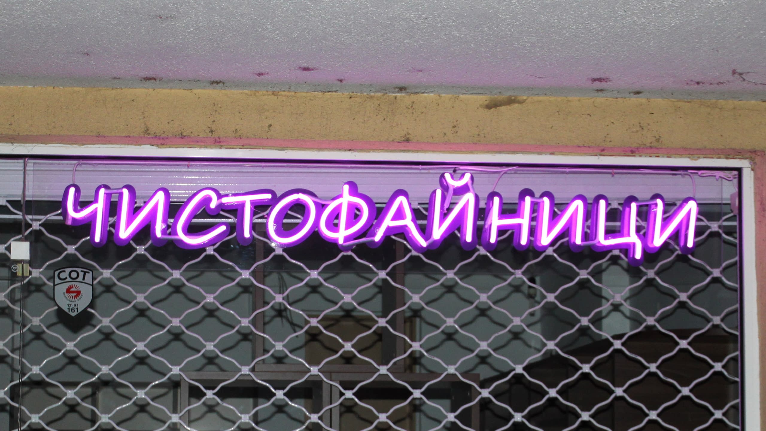 Монтиран лилав светещ надпис зад метална охранителна решетка на витрина на магазин.