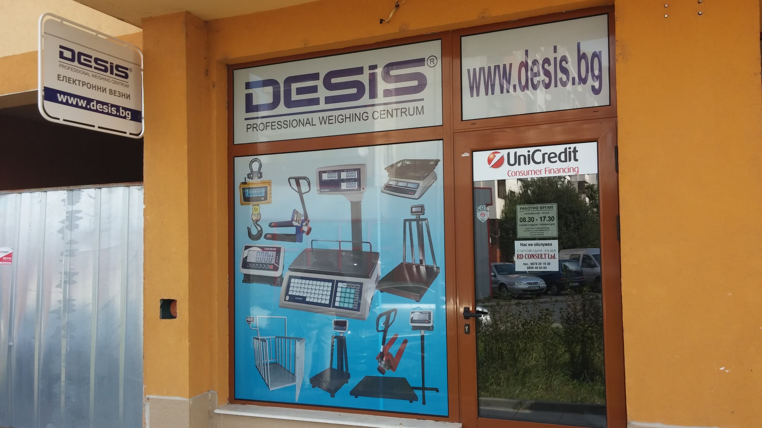 Челен изглед към входа на офис на фирма DESIS, чиято витрина е покрита с фолио с продуктови изображения.