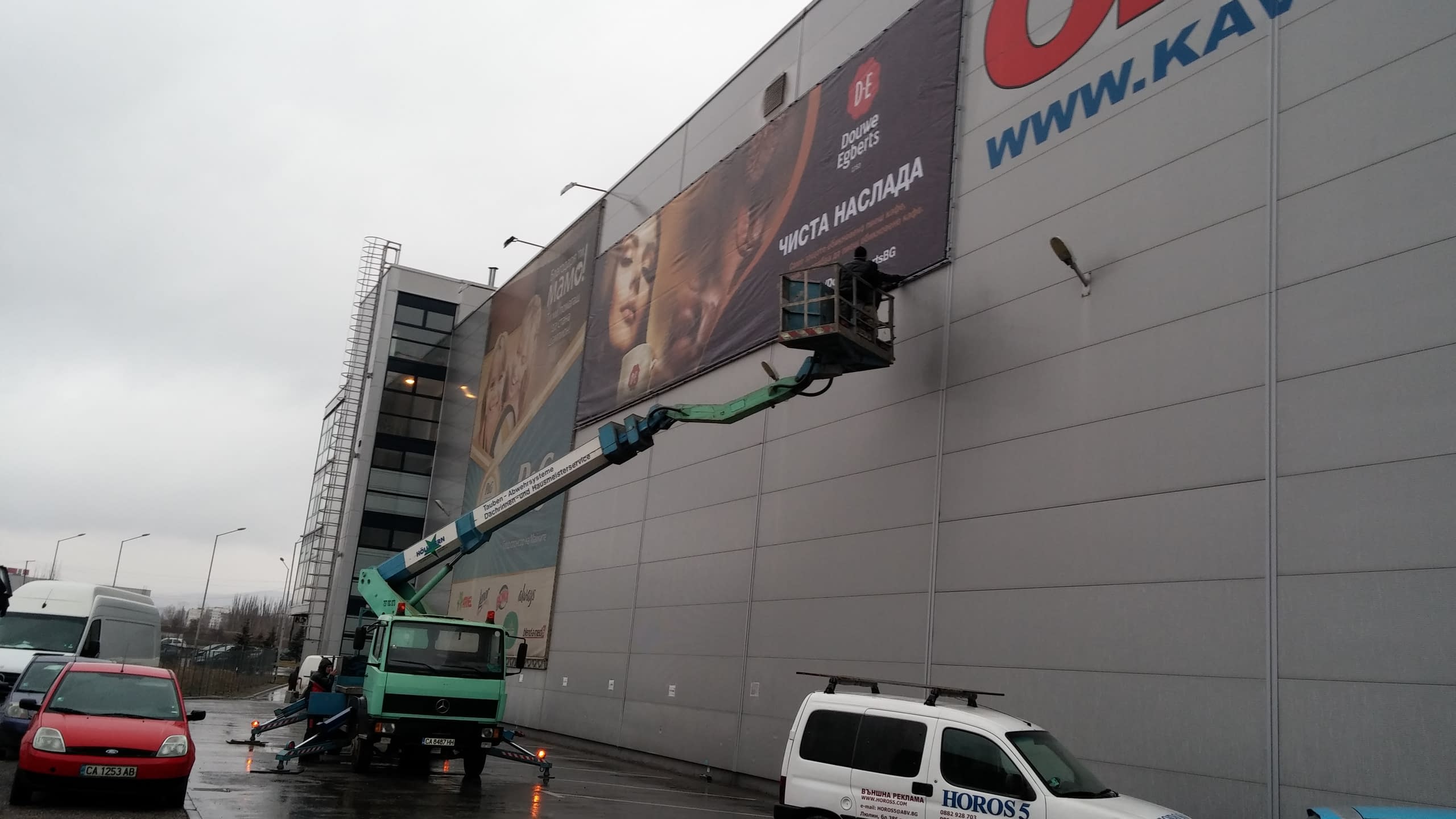 Работник в кош на автовишка монтира голям хоризонтален банер на фасадата на сграда.