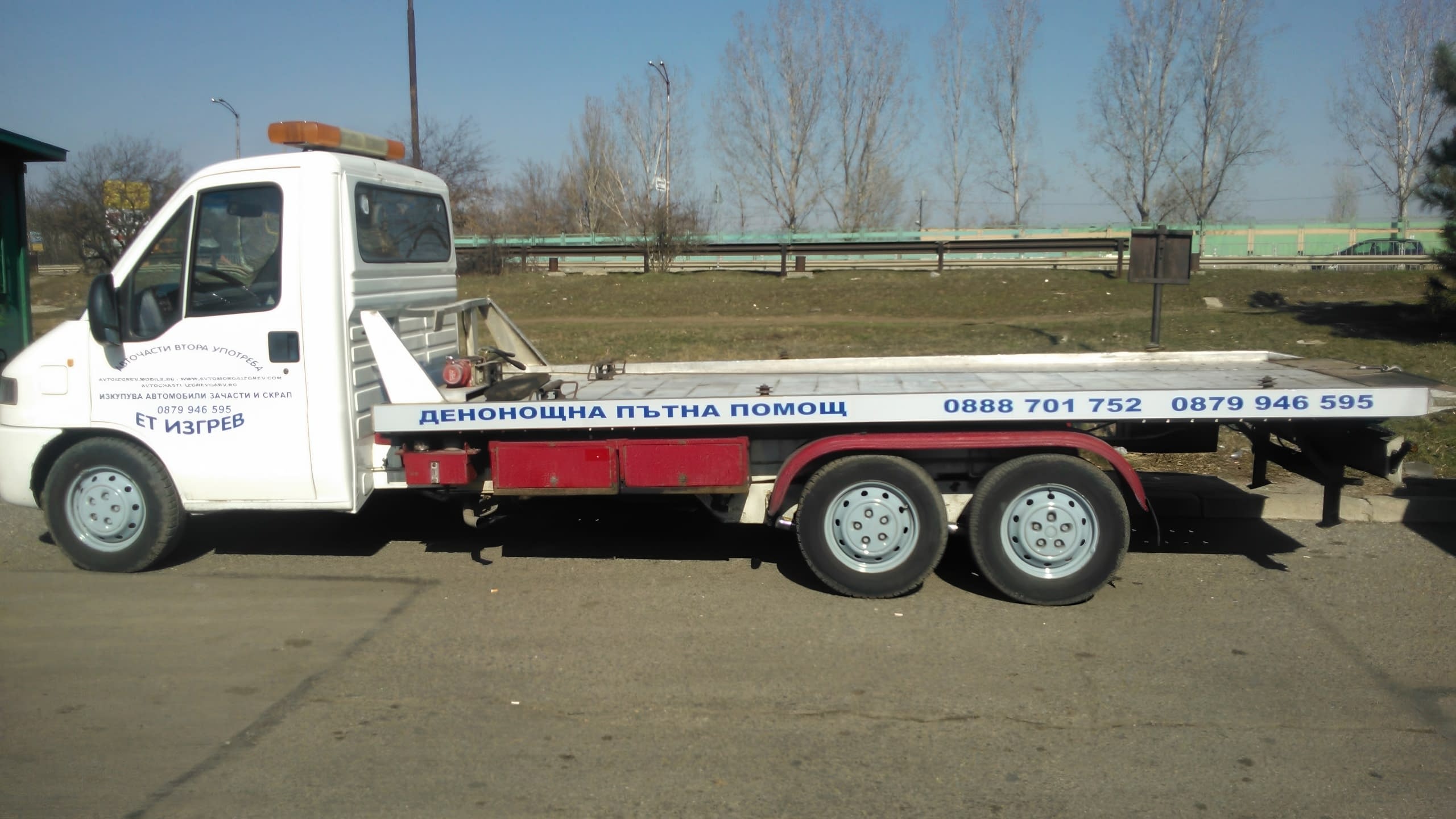 Страничен изглед на бял камион за пътна помощ с надписи върху кабината и платформата.