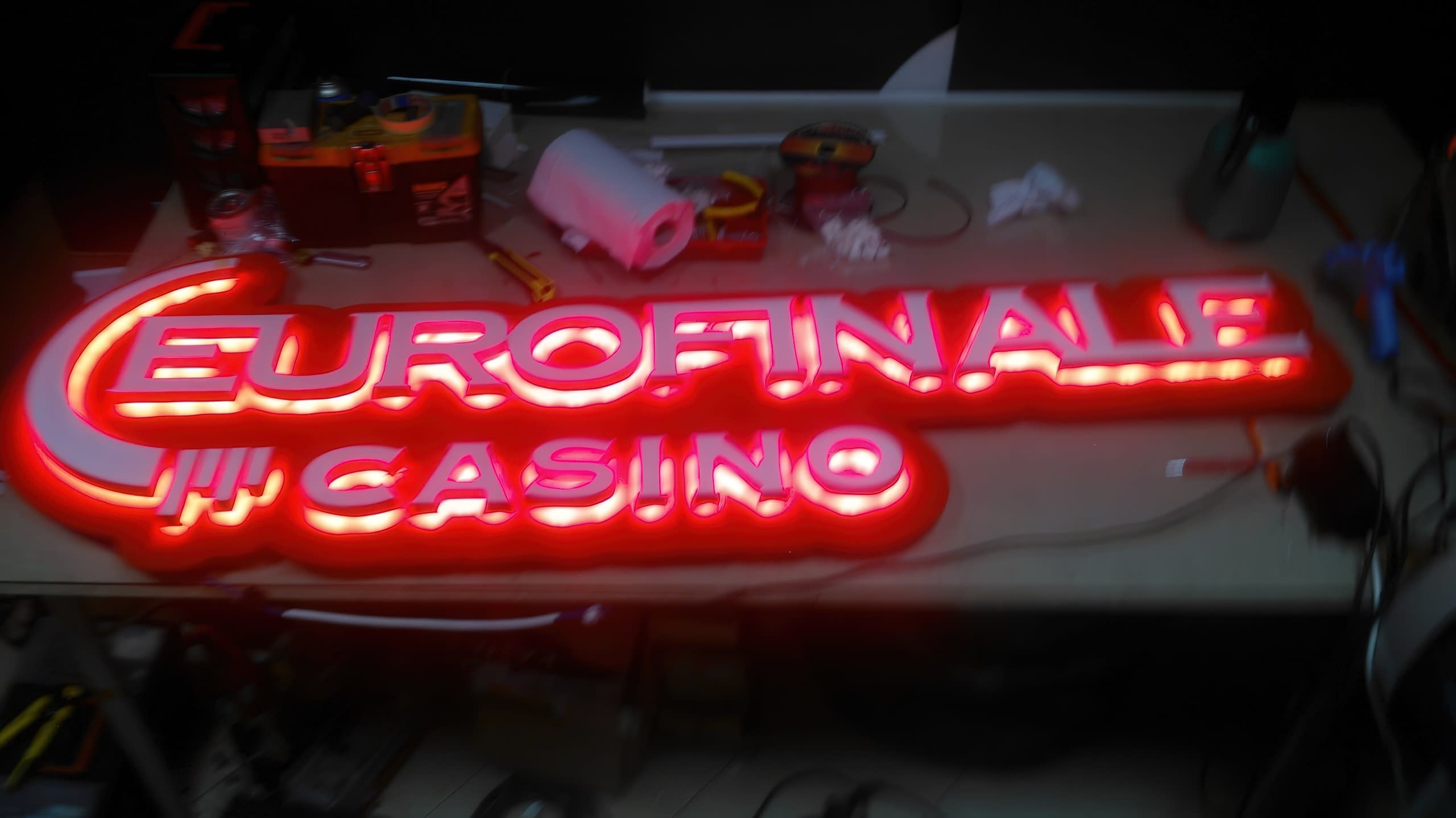 Светеща в червено табела с надпис „EUROFINALE CASINO“, поставена на работна маса в работилница.
