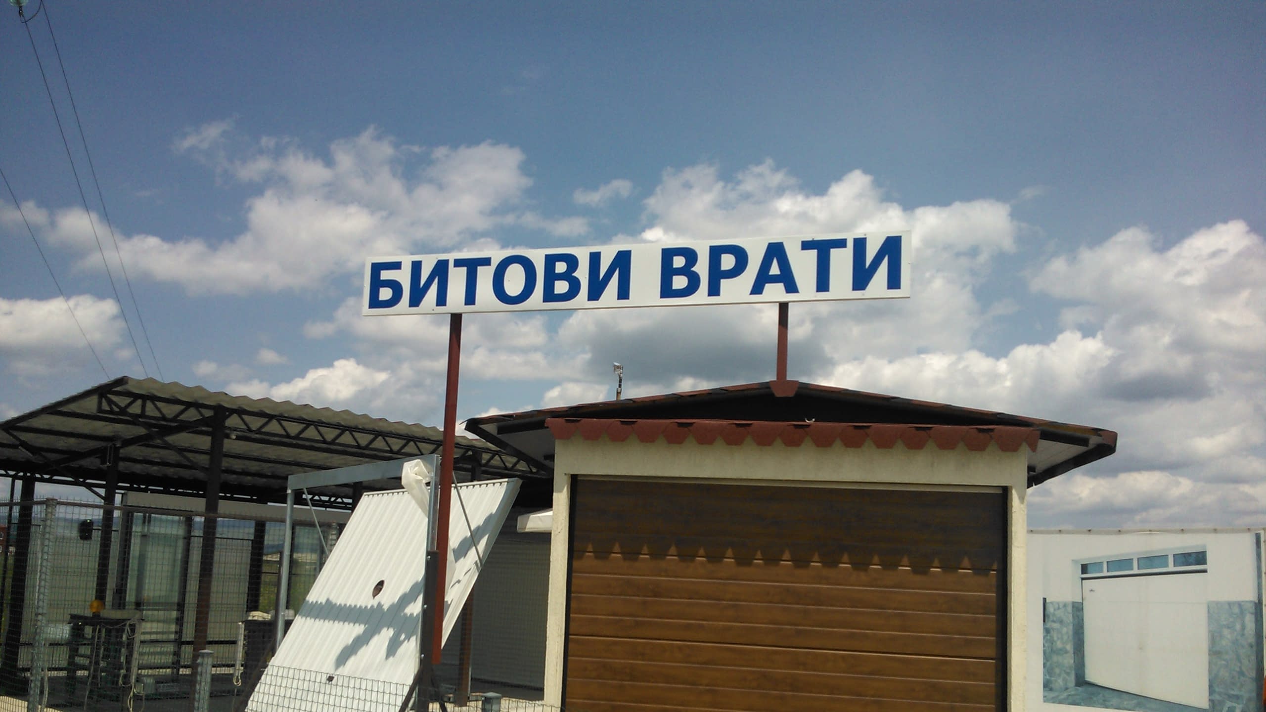 Бяла табела със син надпис на кирилица, монтирана на червени стълбове над покрива на малка постройка с кафява врата.